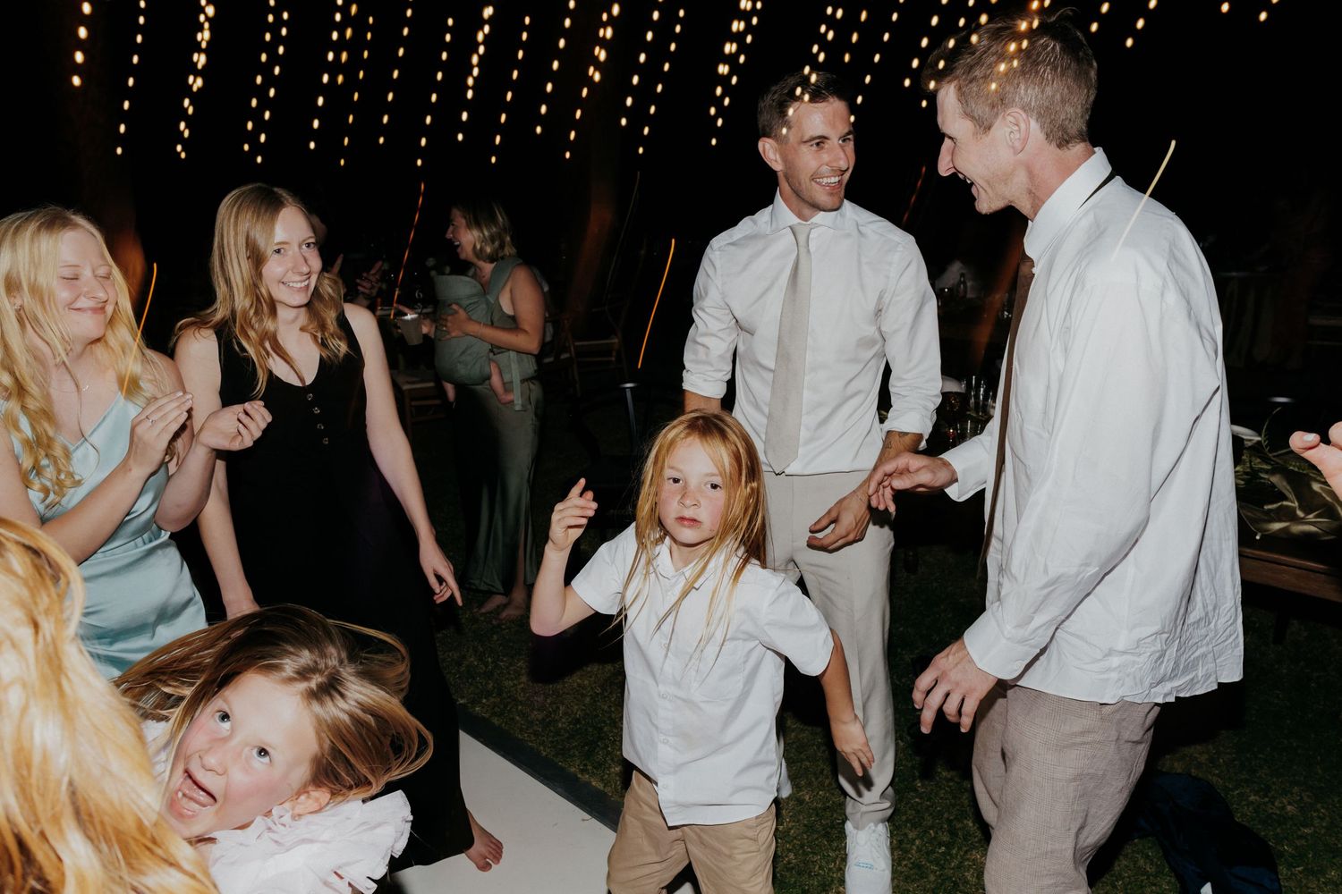 People dancing and celebrating at a nighttime outdoor party under string lights.