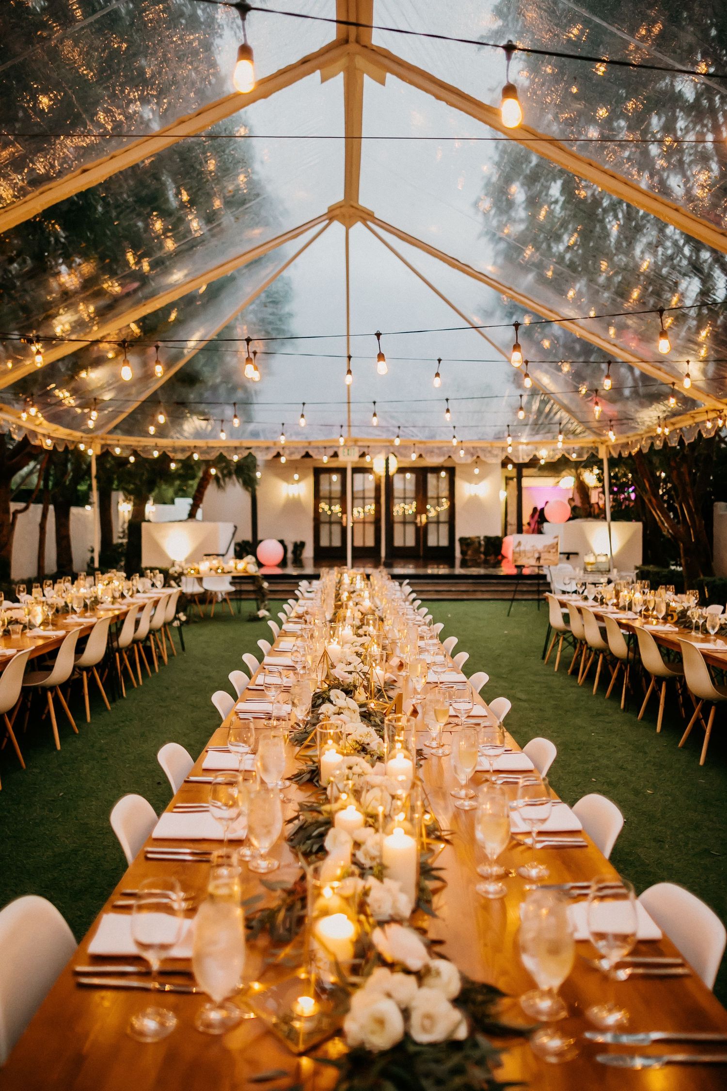 Long banquet tables elegantly set under a clear tent with twinkling string lights create a romantic evening reception atmosphere.