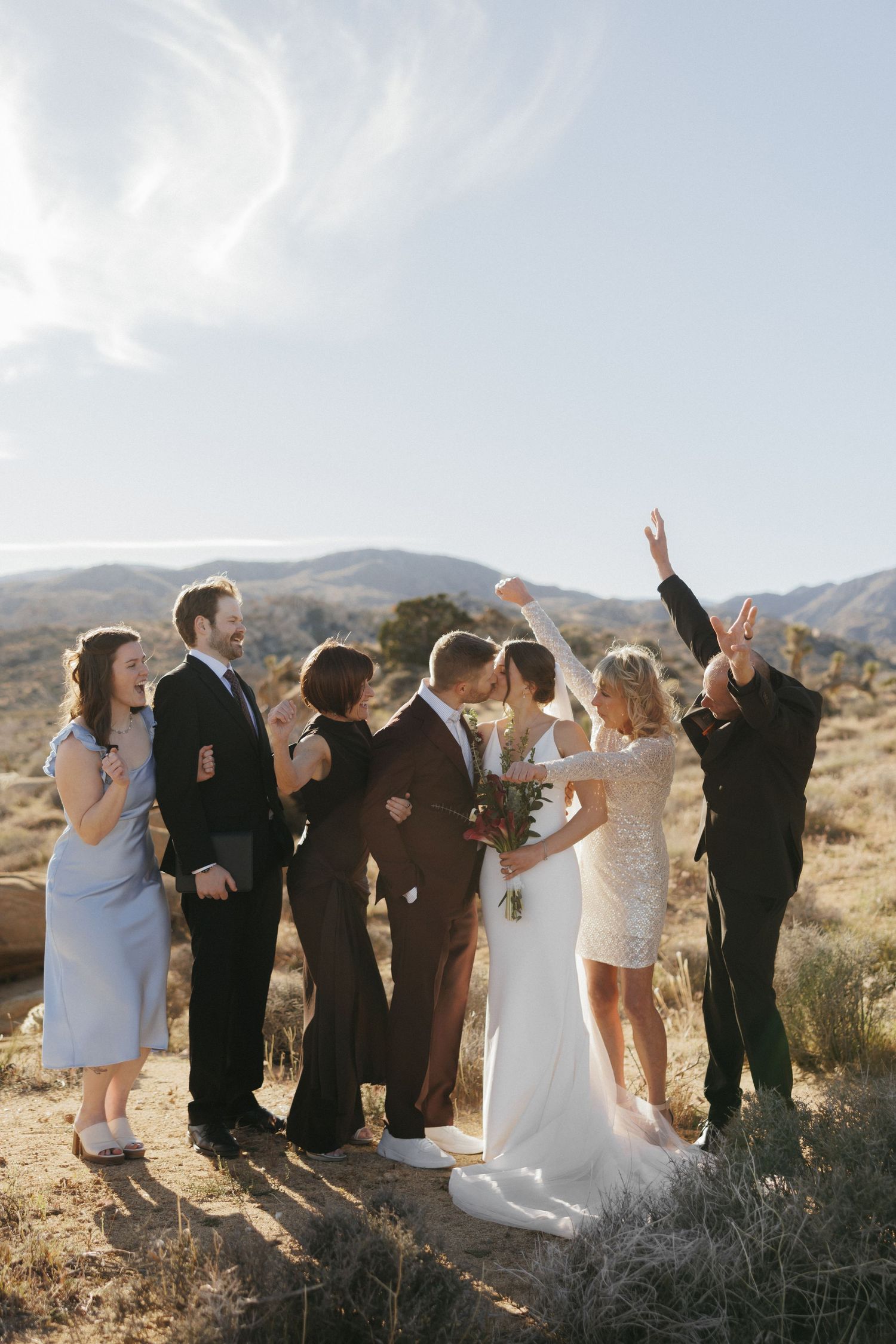 Wedding party celebrates with kiss and cheers in scenic desert landscape.