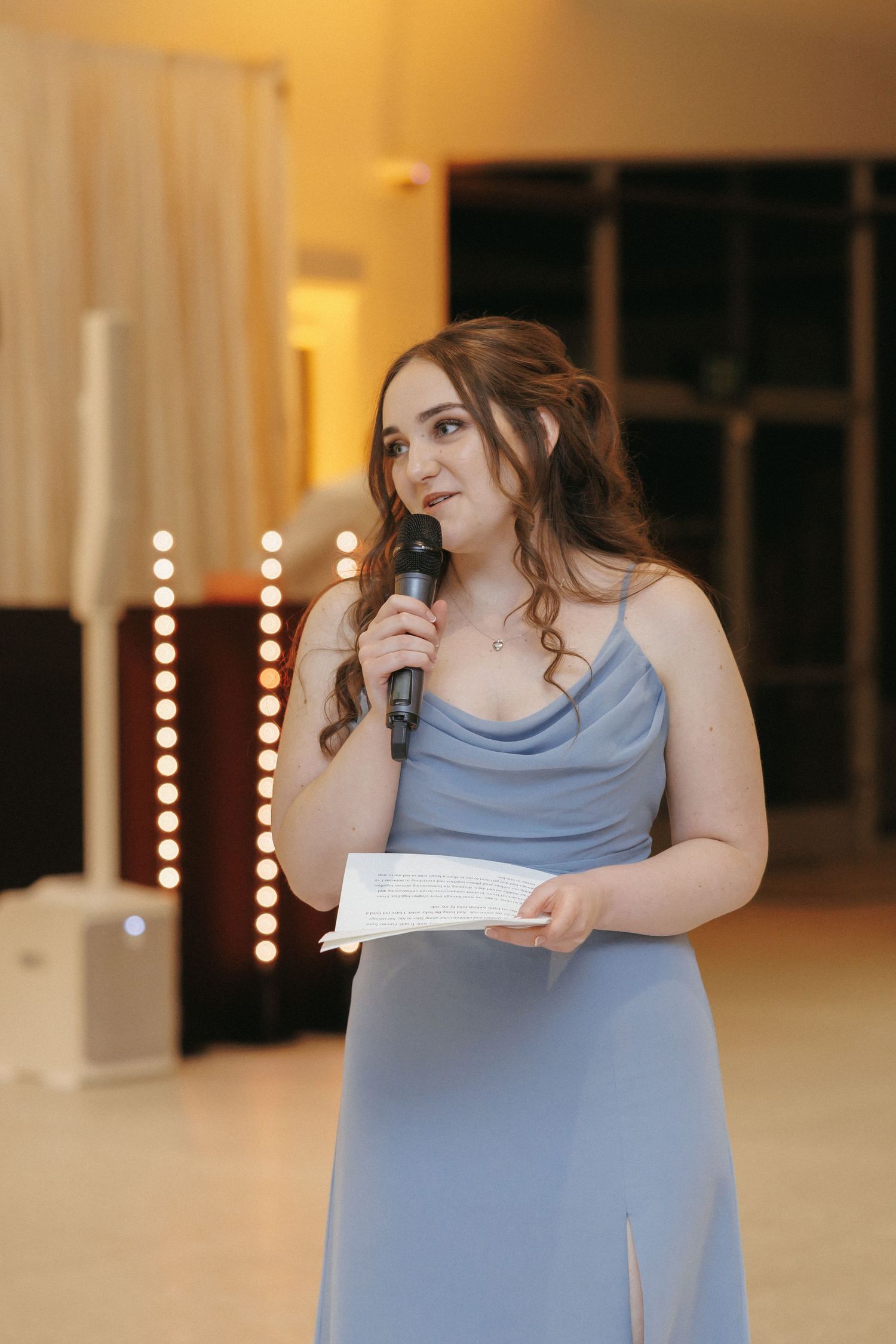 Bridesmaid in blue dress delivers heartfelt speech holding microphone and notes at wedding reception.