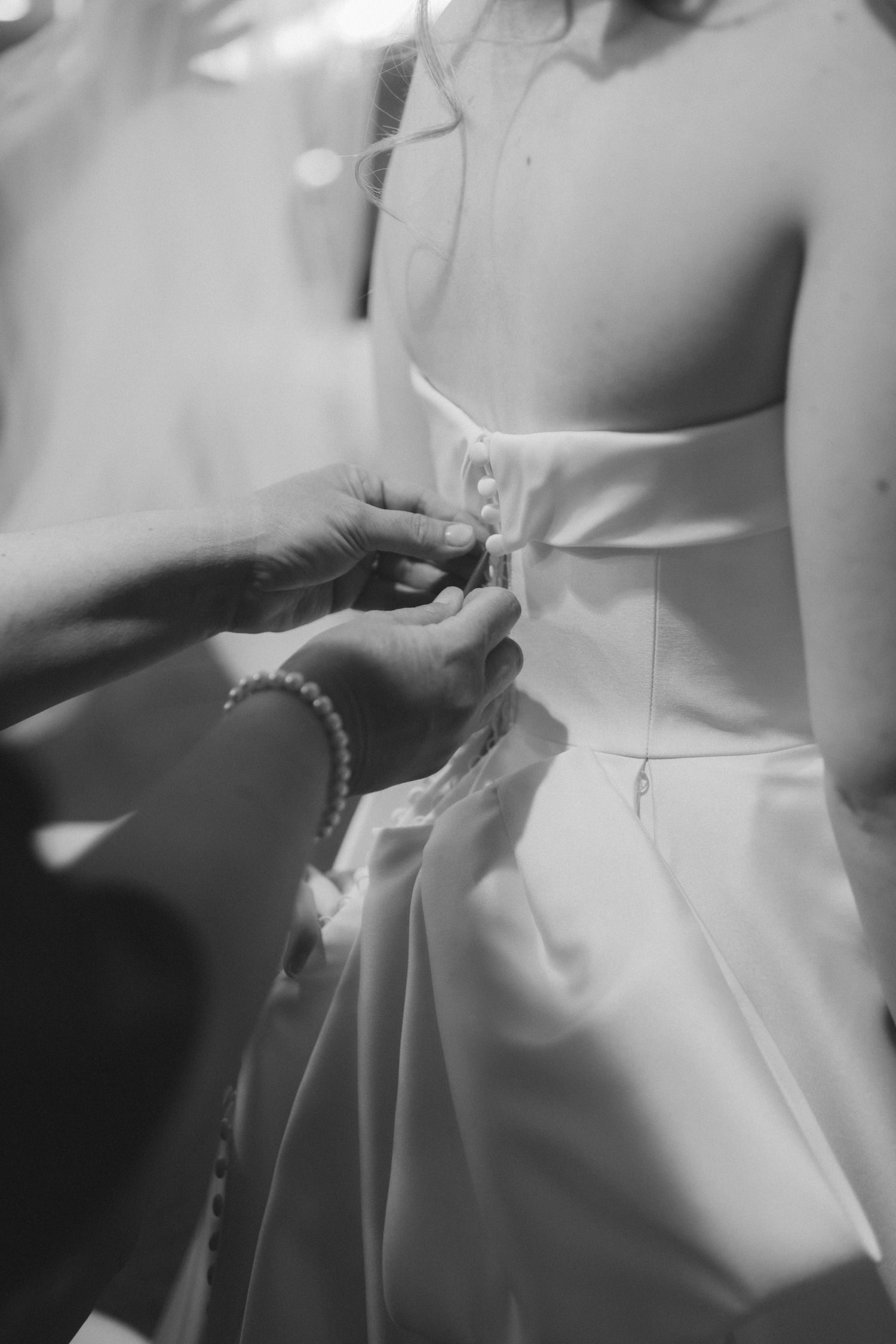 Black and white photo of hands buttoning the back of a white wedding dress.