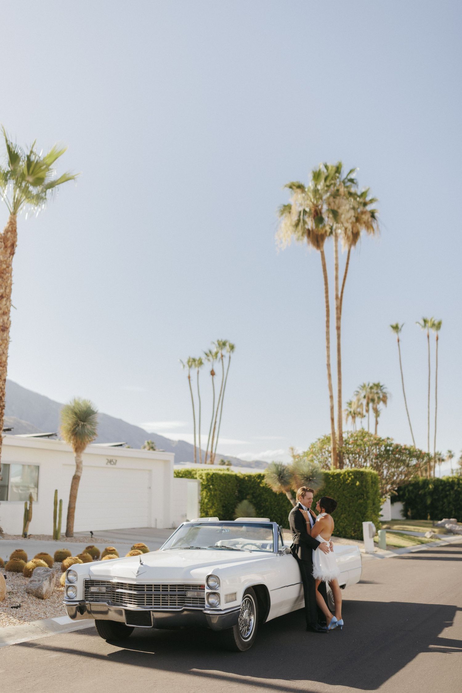 Bride and groom embrace beside a vintage white Cadillac convertible surrounded by palm trees in Palm Springs.