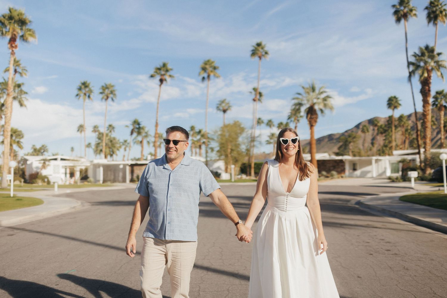 Couple holding hands walking down a sunny Palm Springs street lined with palm trees and mid-century modern homes.