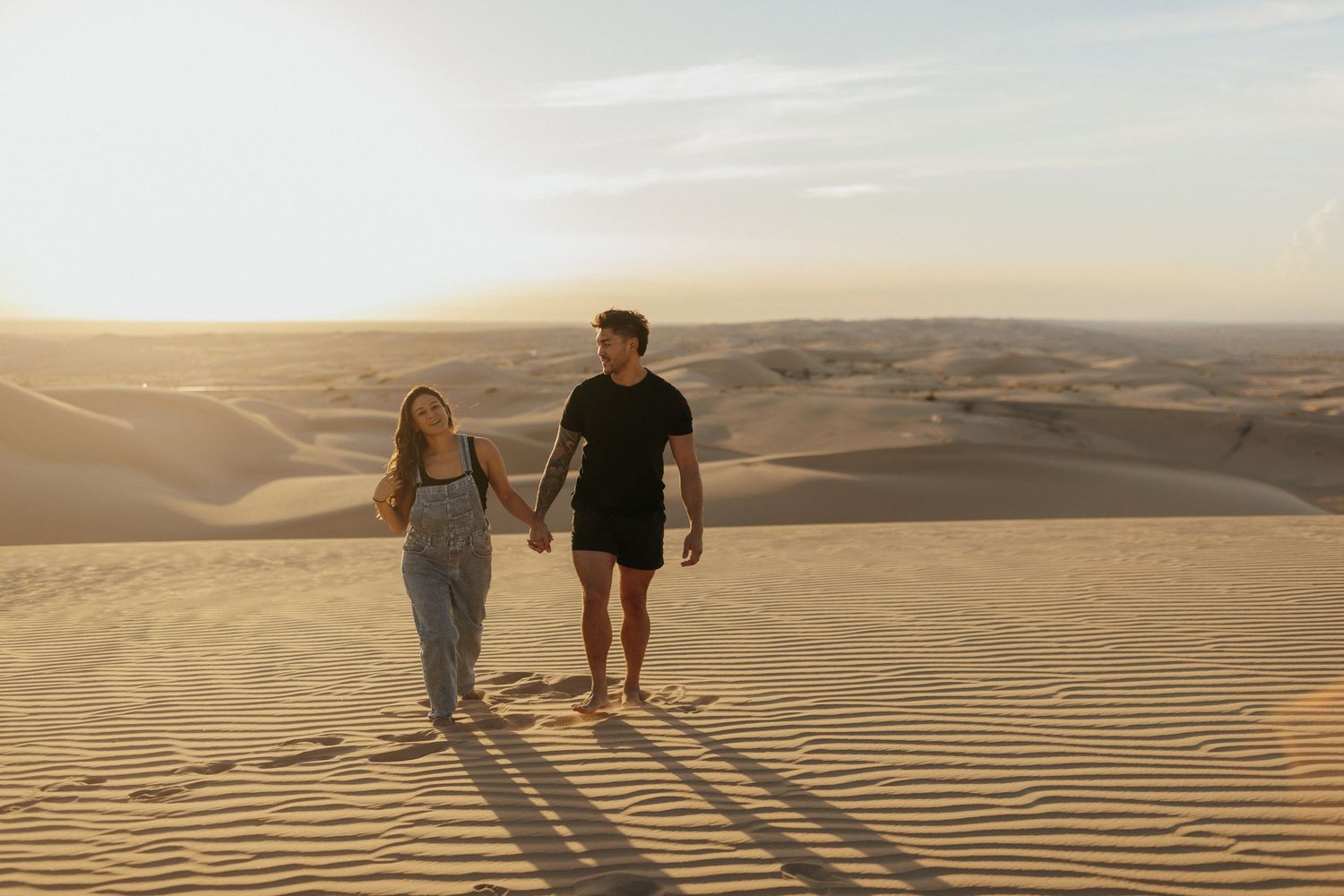 Two figures walking across scenic desert sand dunes at sunset with long shadows stretching across rippled sand.