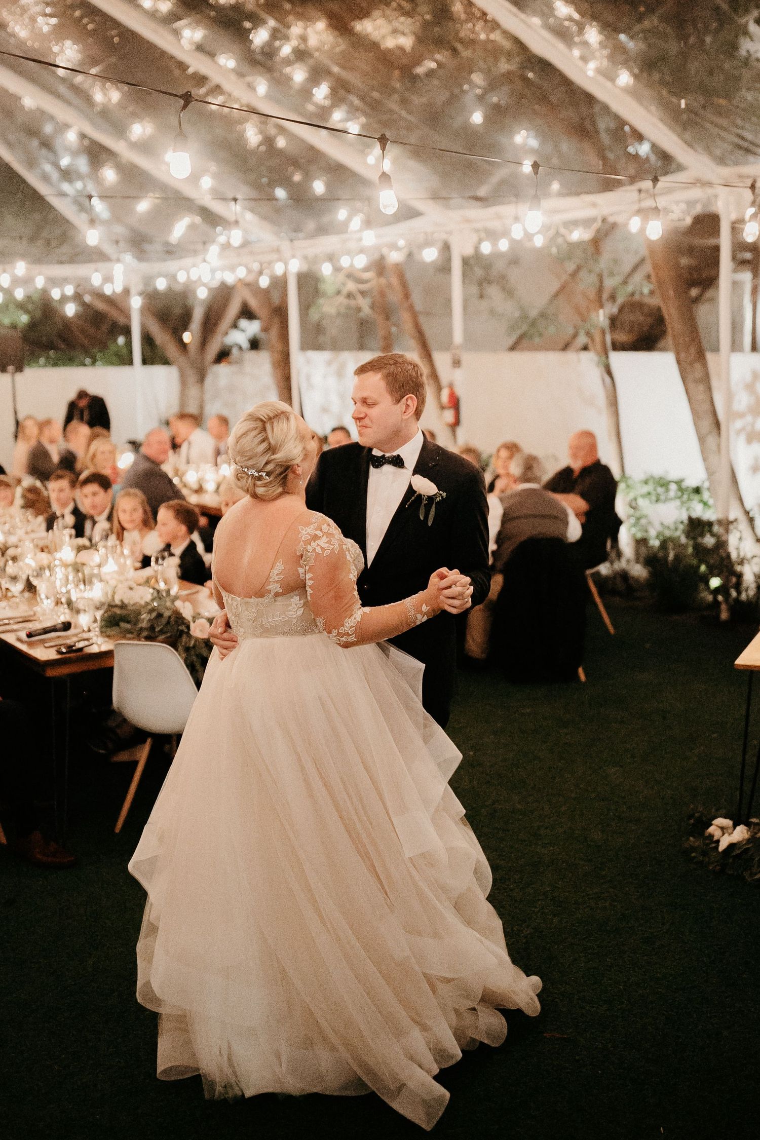 A romantic first dance captured under a tent with soft string lights as wedding guests look on at an evening reception.