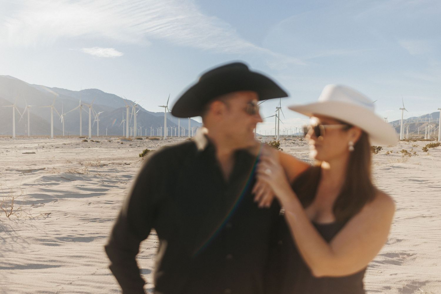 Couple in cowboy hats sharing a romantic moment in a desert landscape with wind turbines and mountains behind them.