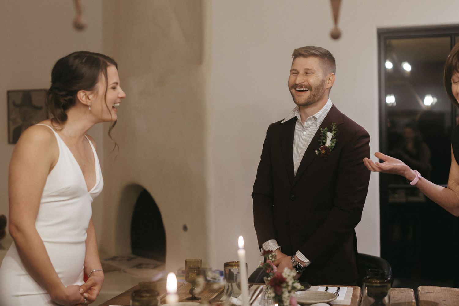 Bride and groom laugh joyfully together at candlelit wedding reception dinner table indoors.