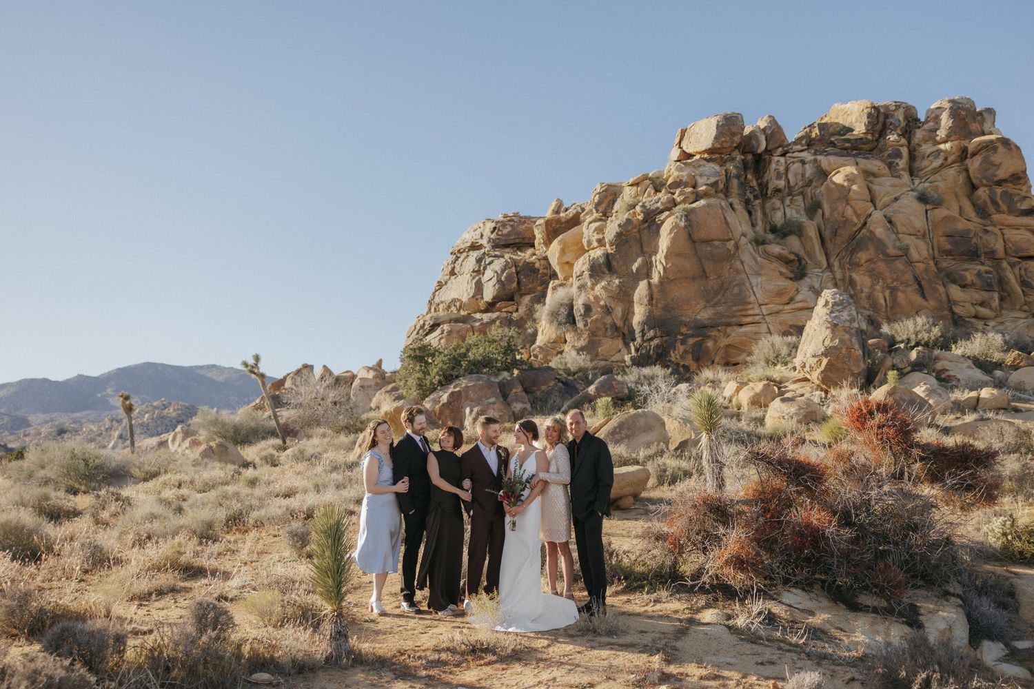 Small wedding ceremony group gathered before massive boulder formations in Joshua Tree desert landscape.
