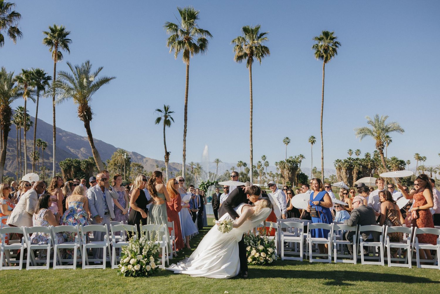 Bride and groom sharing first kiss at outdoor Palm Springs wedding ceremony with palm trees.