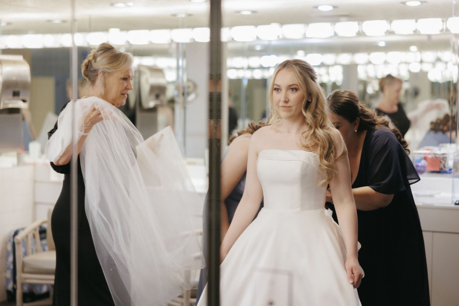 Bride in white strapless gown with veil being adjusted by assistant in dressing room with Hollywood mirrors.