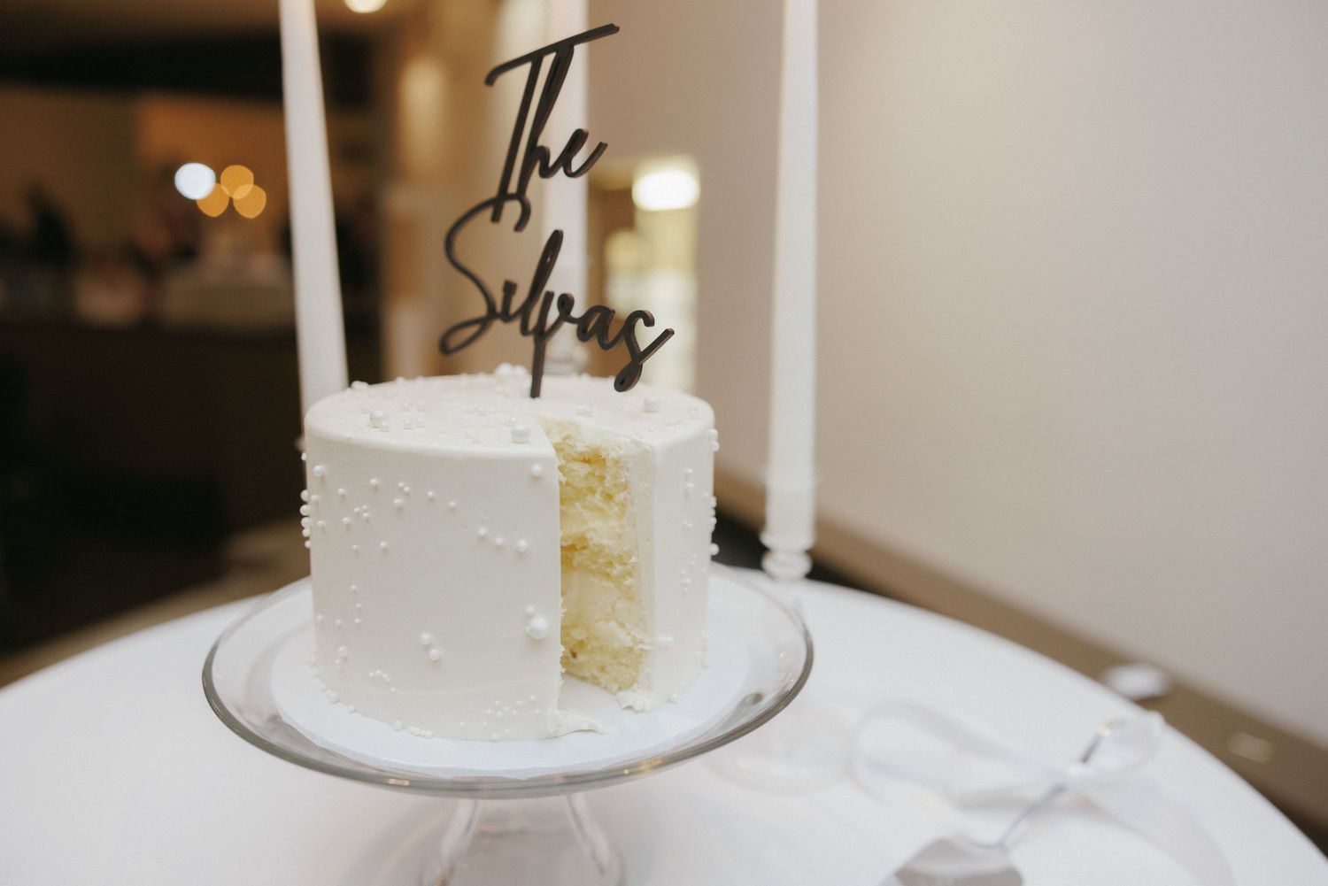 Single-tier white pearl wedding cake with The Silvas cake topper on glass stand, partially sliced.