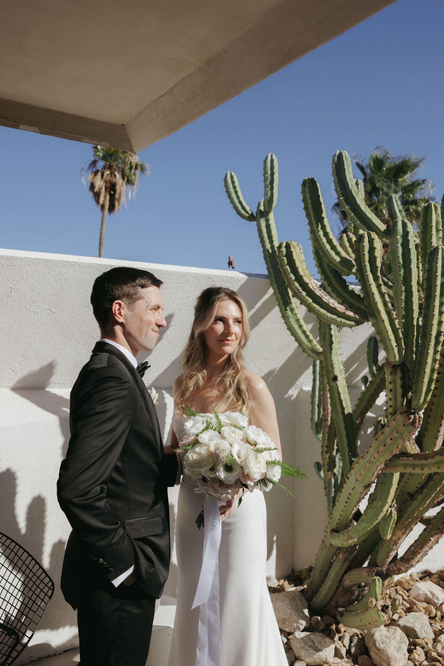 Bride and groom stand beside a tall cactus against a white stucco wall in a sunny desert wedding setting.