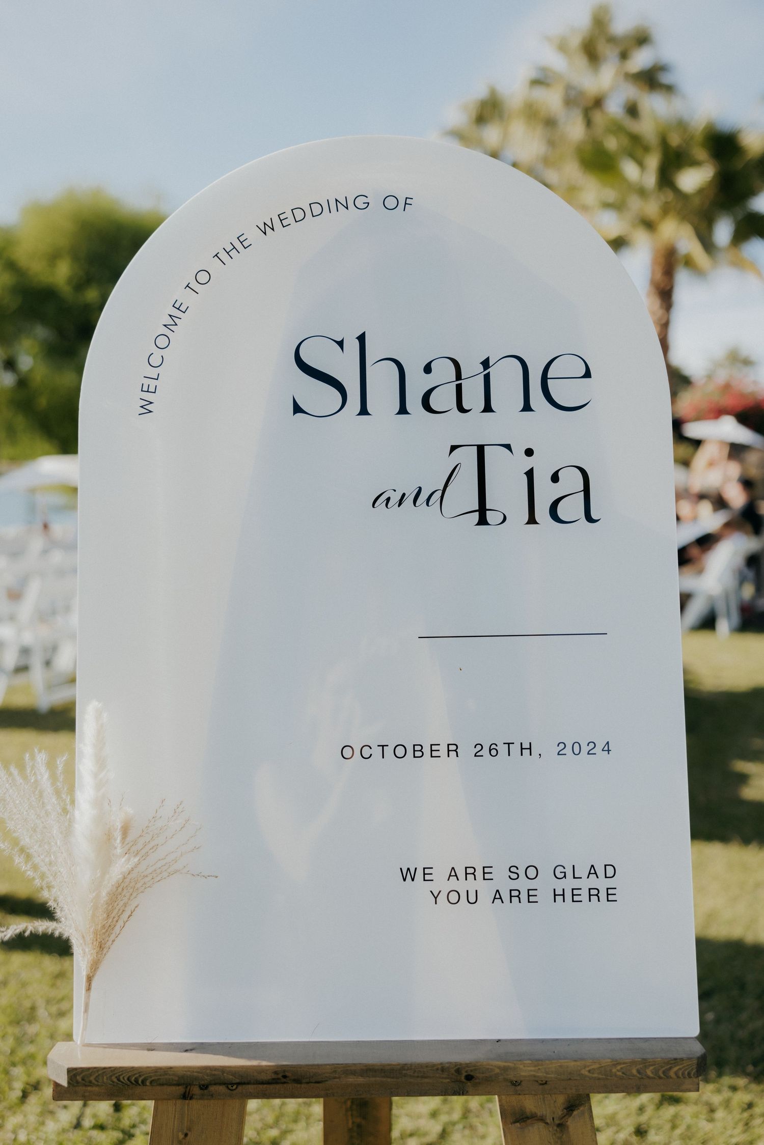 Elegant white curved wedding sign displaying 'Shane and Tia' with text 'October 26th, 2024' in modern typography against outdoor setting.