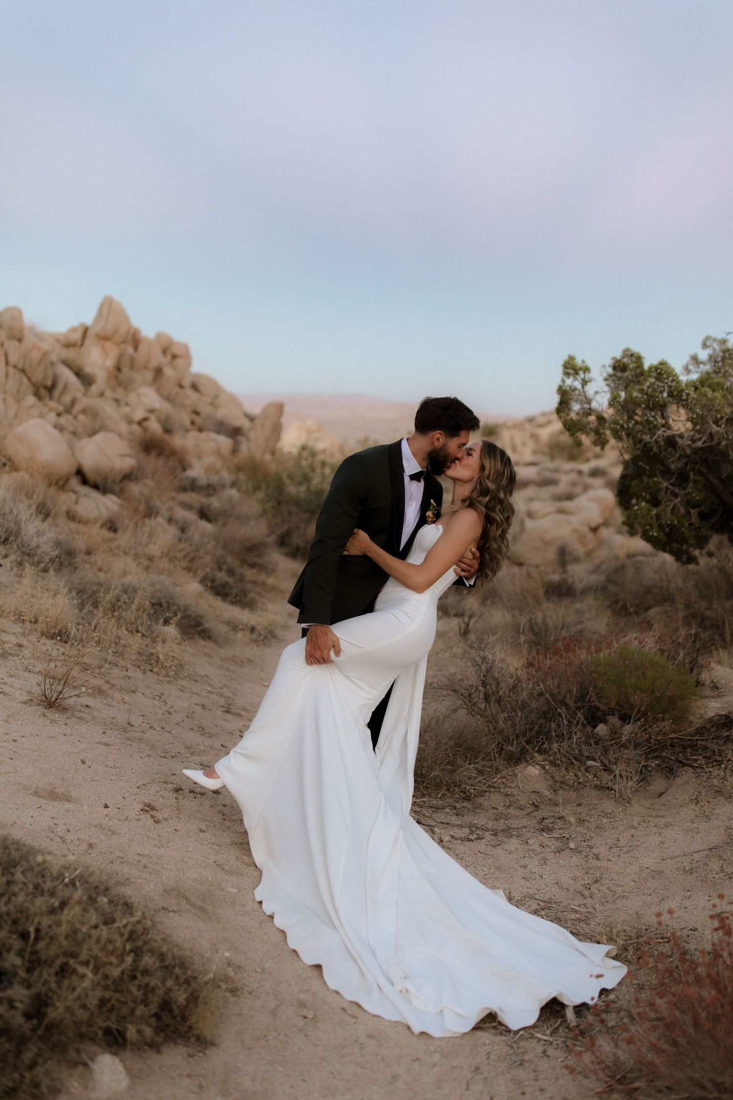 Couple in wedding attire embrace on a desert trail surrounded by rocky terrain at dusk.