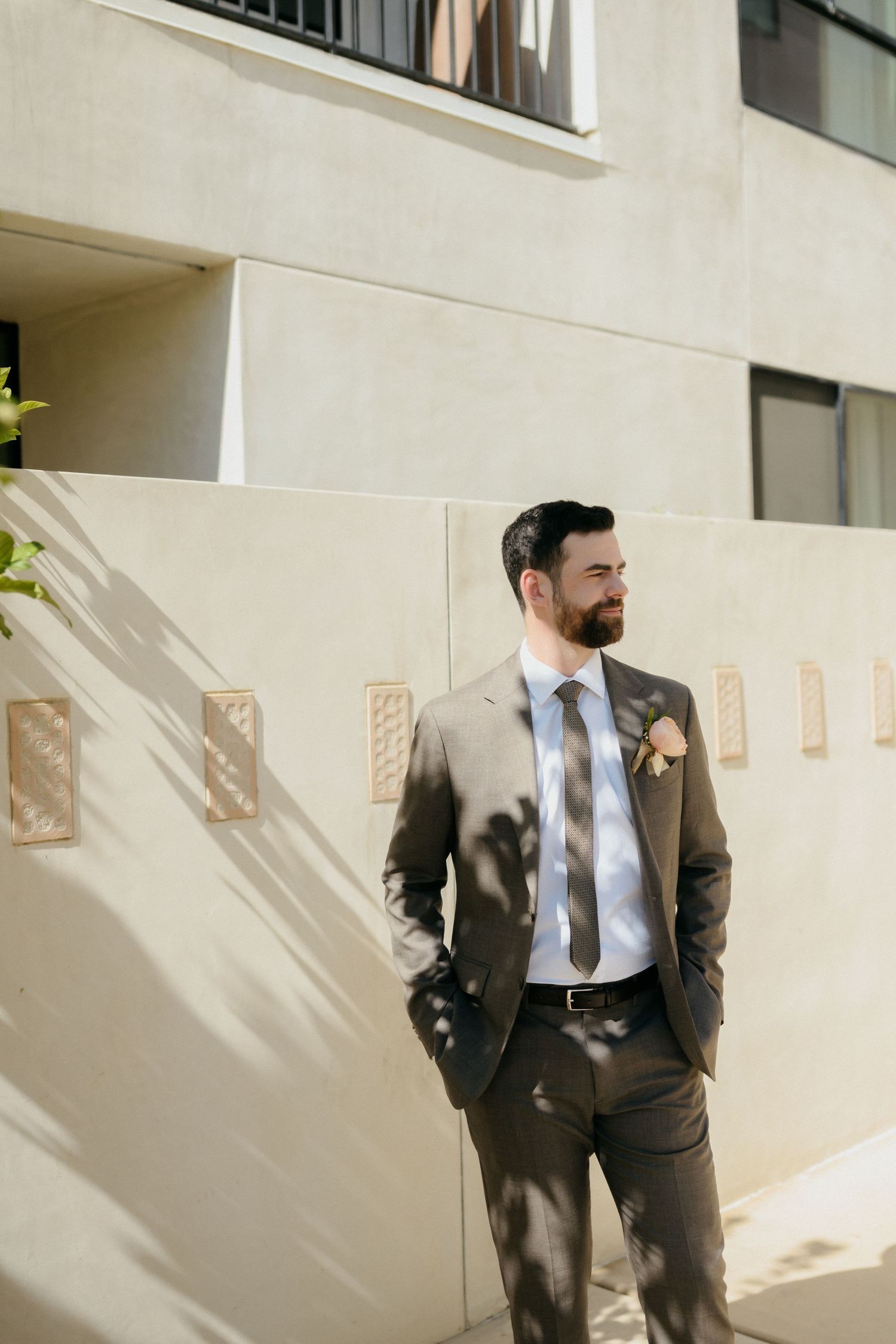 A groom in a brown suit and blue tie stands confidently against a modern white exterior wall in natural sunlight.