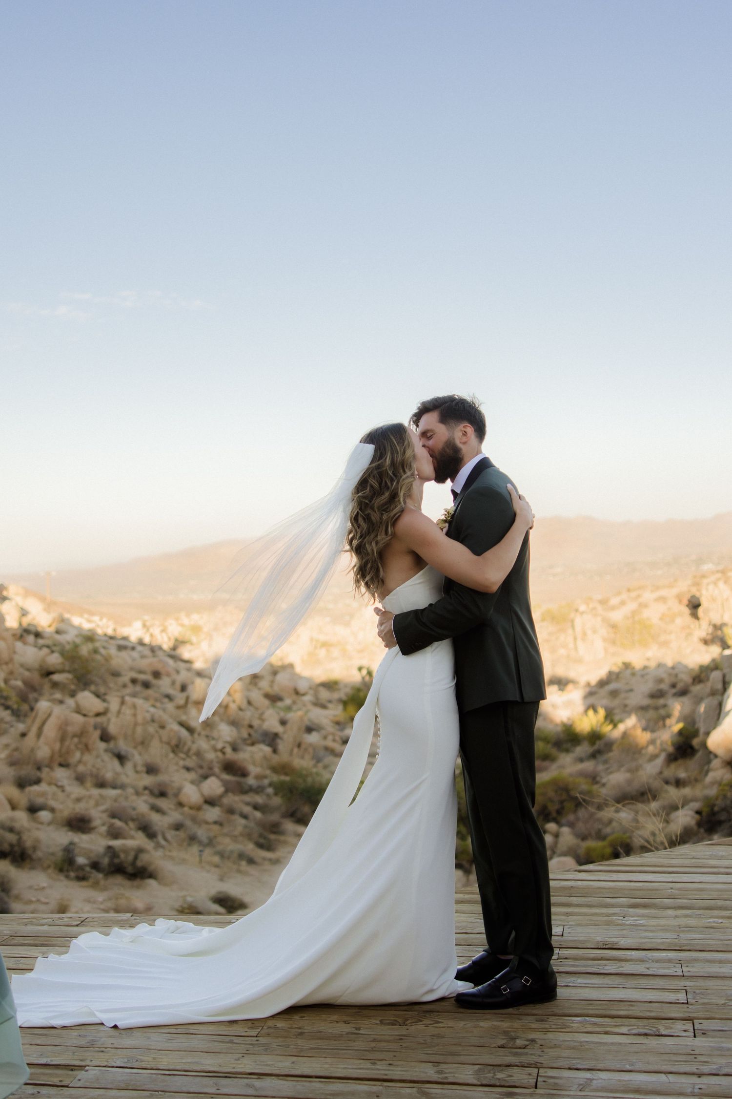 A romantic couple in wedding attire sharing an intimate moment on a wooden deck overlooking a desert landscape at sunset.