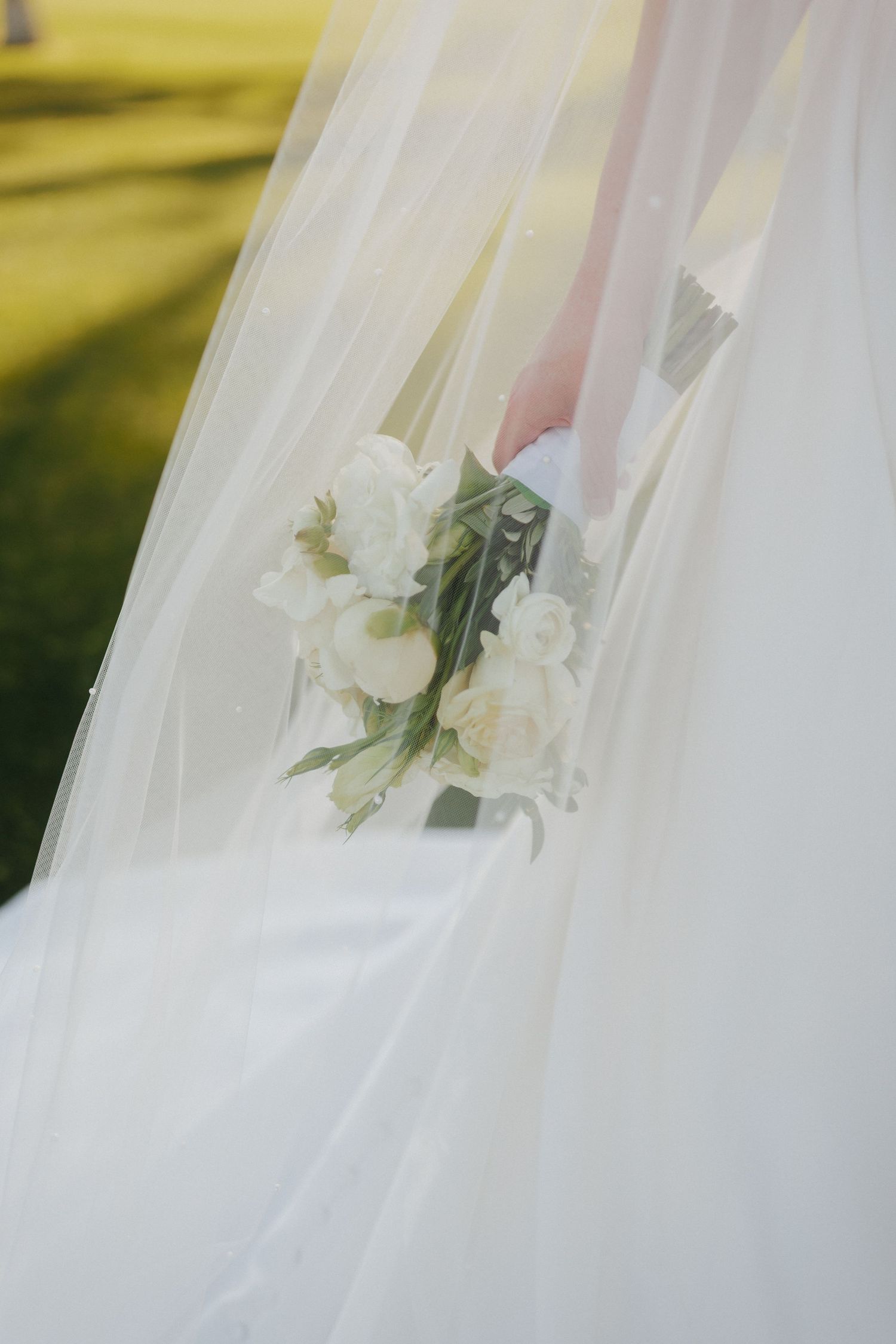 Bride holding small white rose bouquet beneath flowing veil in soft natural light.