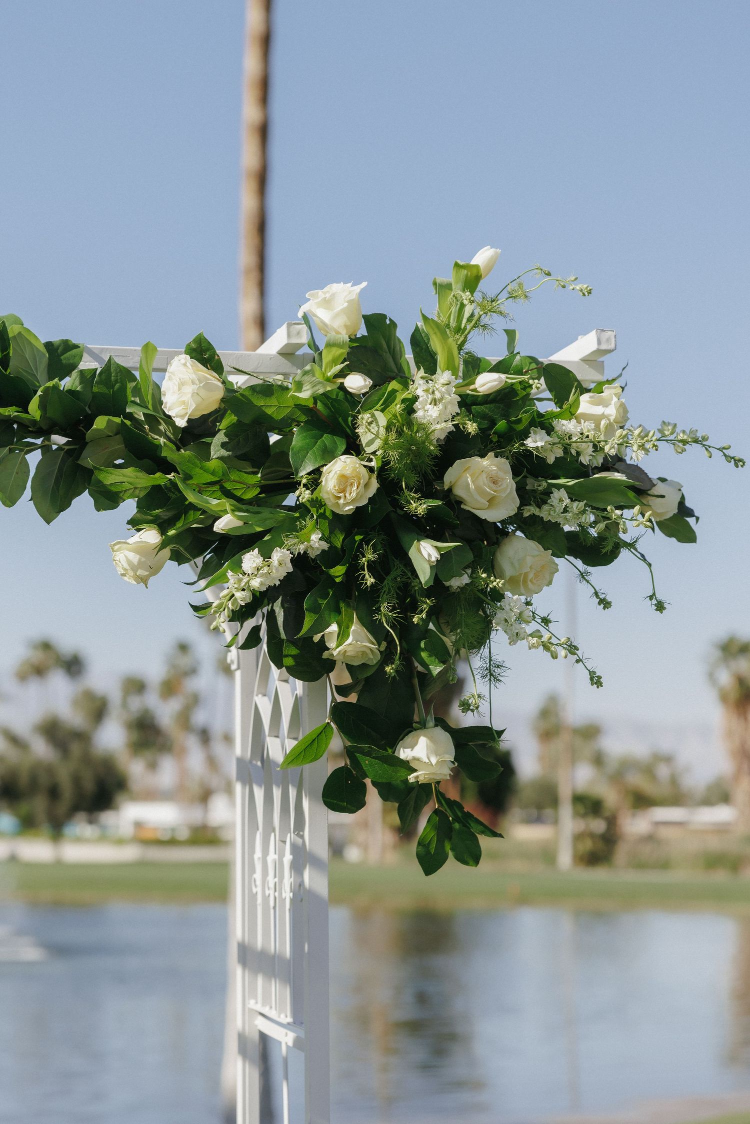 White roses and greenery floral arrangement on a white wedding arch outdoors near a lake.