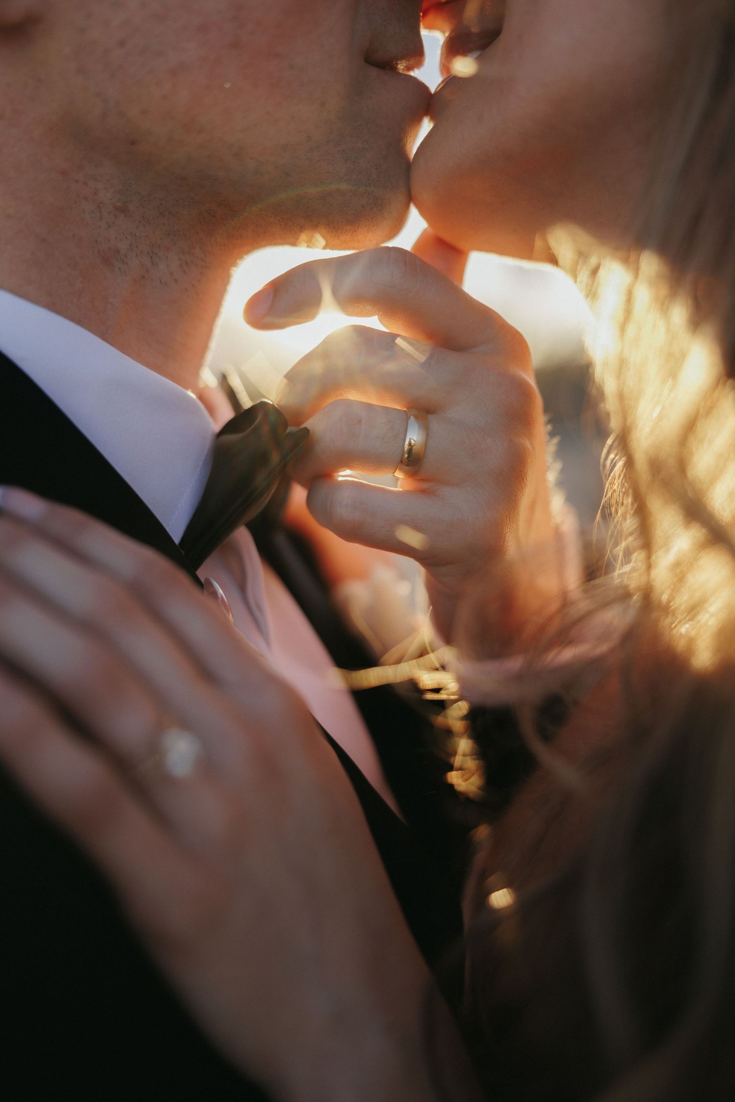 Couple sharing a romantic kiss at golden hour, with wedding rings visible on their intertwined hands.