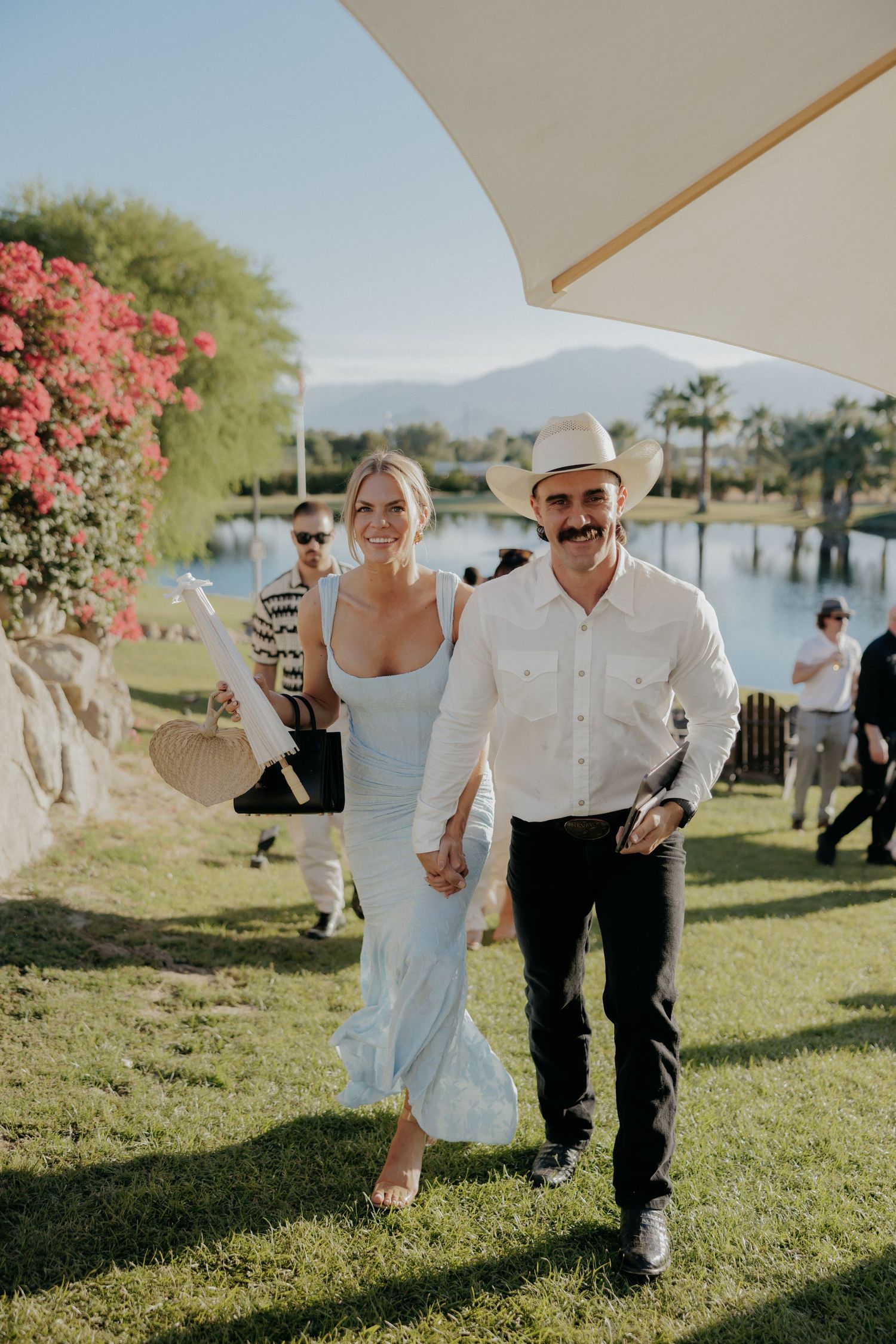 A couple walks on green grass near a lake with pink bougainvillea flowers and mountains in the background.