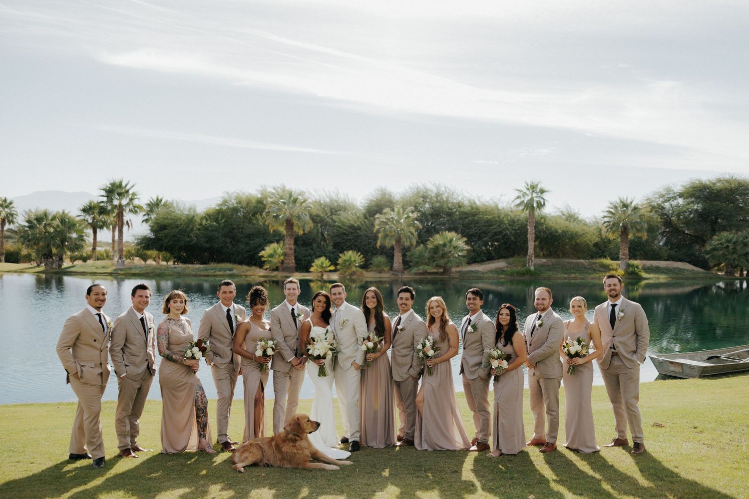Panoramic view of wedding party in matching beige outfits lined up across green lawn for formal photo.