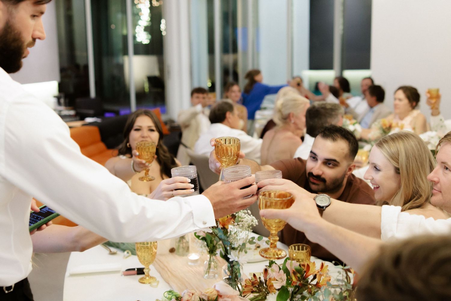 Wedding reception guests at formal dining tables in a light-filled venue decorated with flower centerpieces and linens.