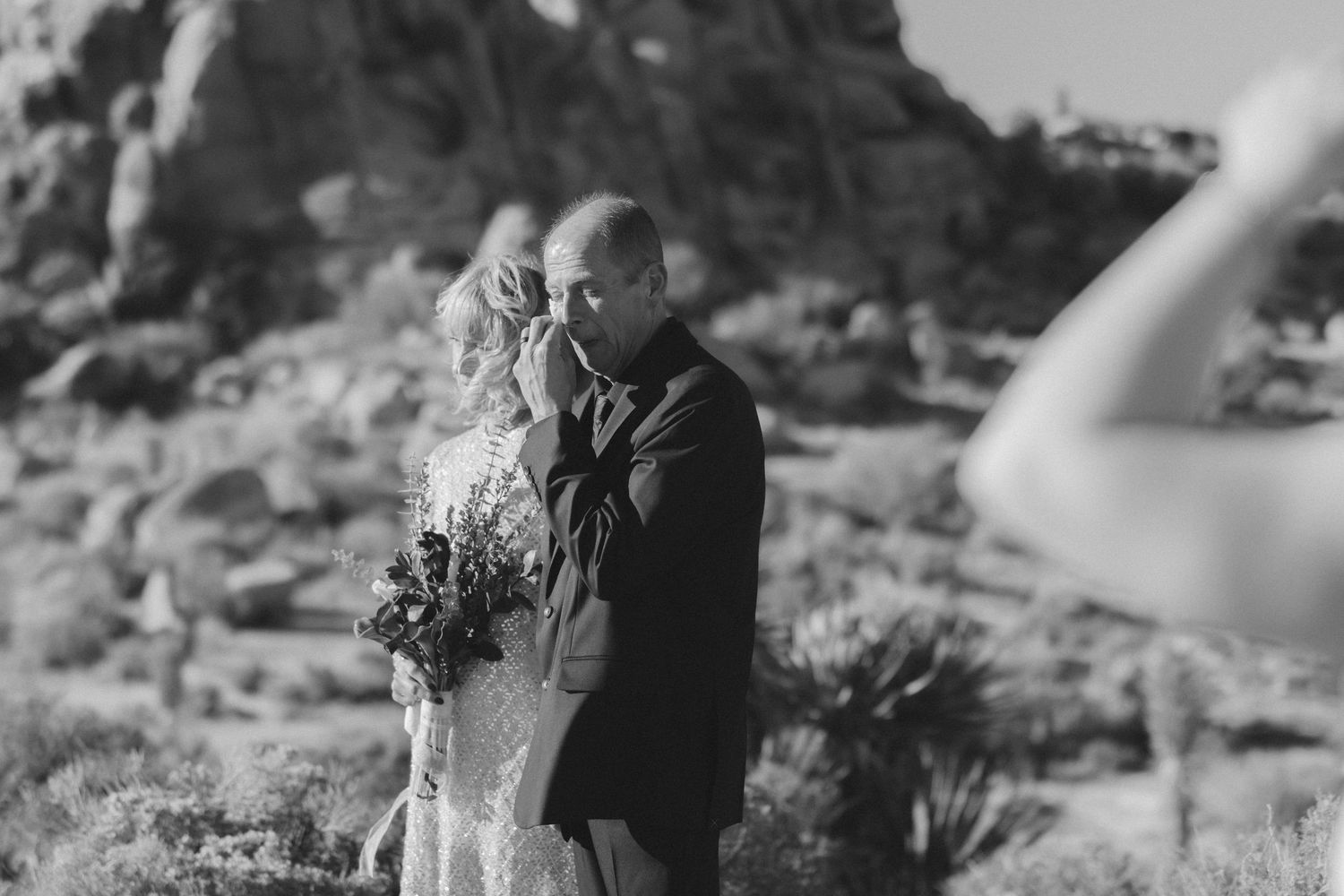 Groom kissing bride's hand in a romantic black and white desert wedding photo with rocky landscape backdrop.