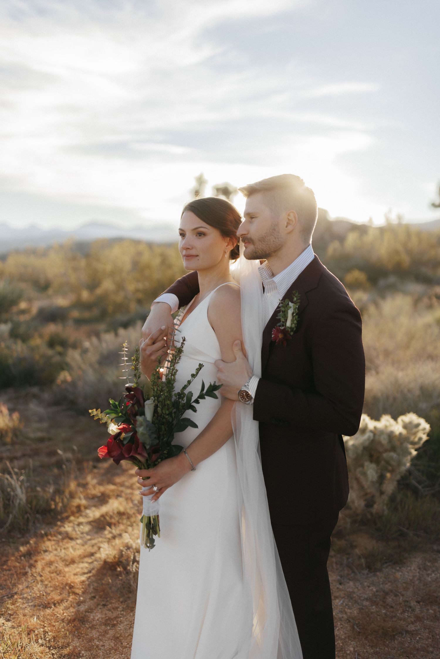 Bride holding red bouquet with groom embracing her from behind in sunlit desert landscape.