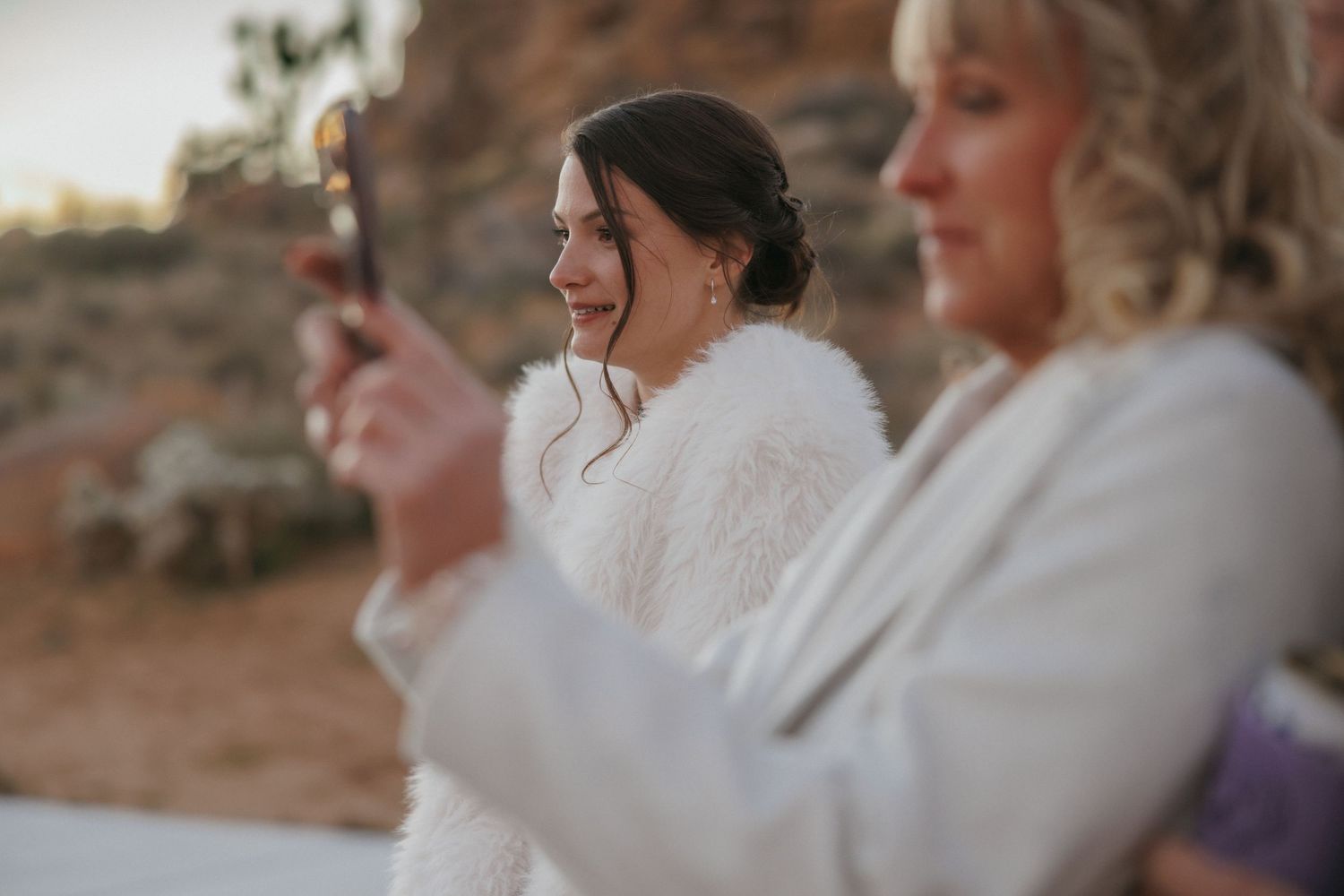 Smiling bride in white fur wrap enjoys a candlelit moment outdoors at desert wedding celebration.