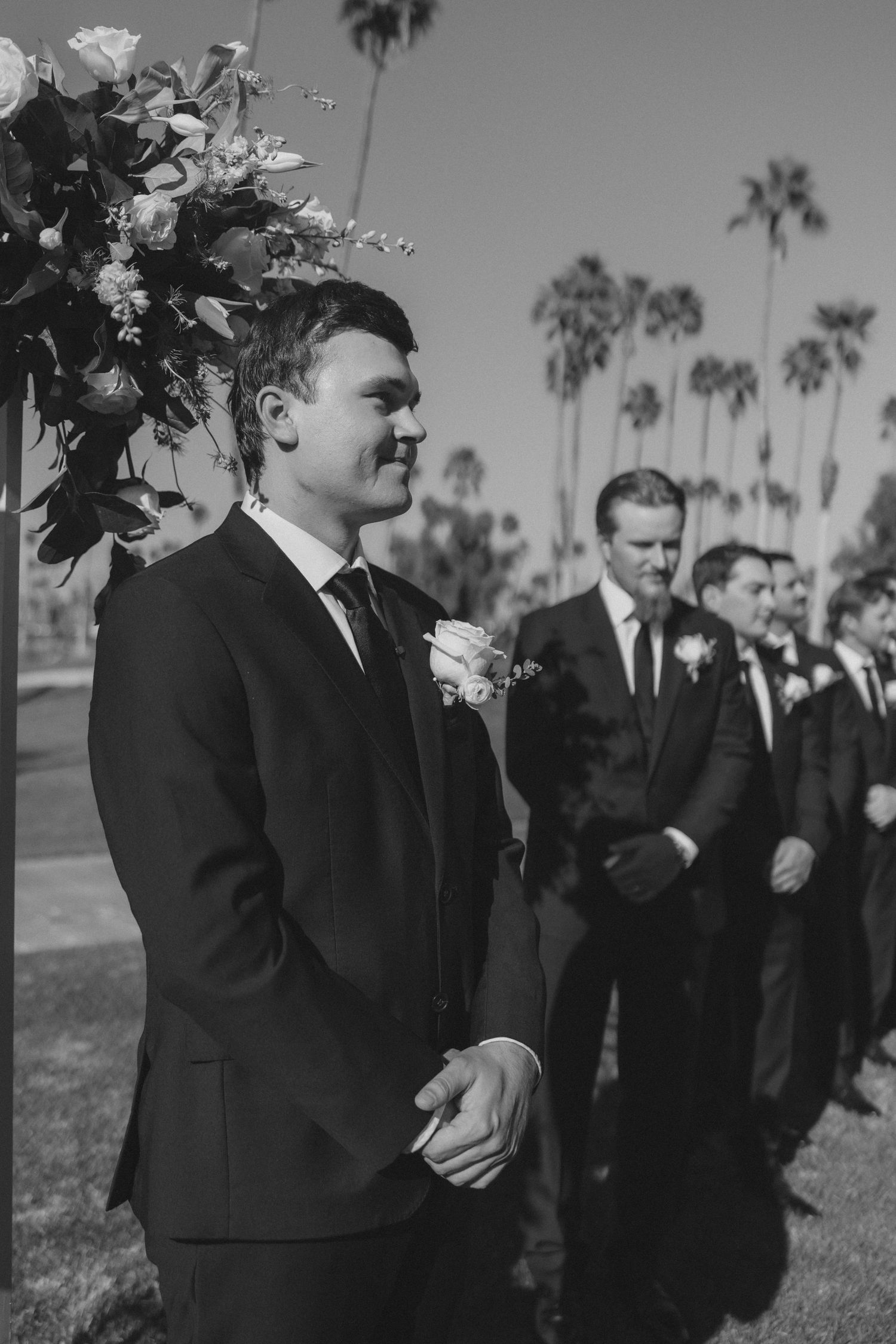 Groom in black suit smiling at altar with groomsmen lined up behind him at outdoor palm tree wedding.