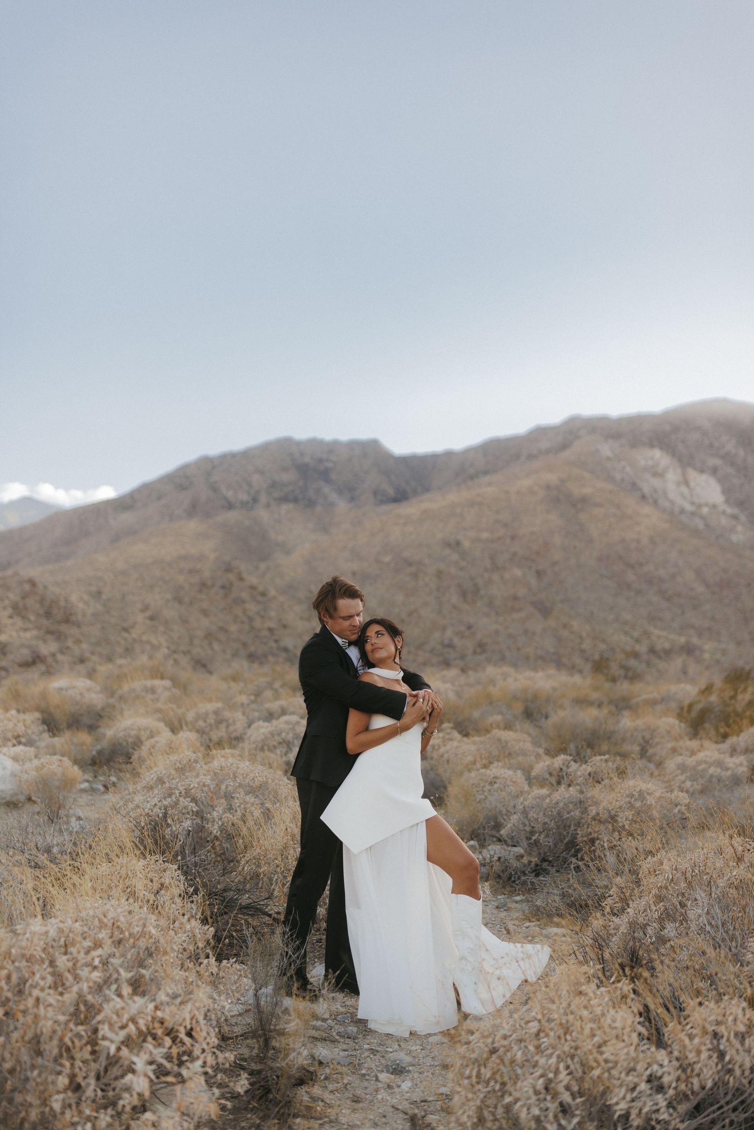 Groom embraces bride in white gown amid desert scrubland with rugged mountains under a pale blue sky.