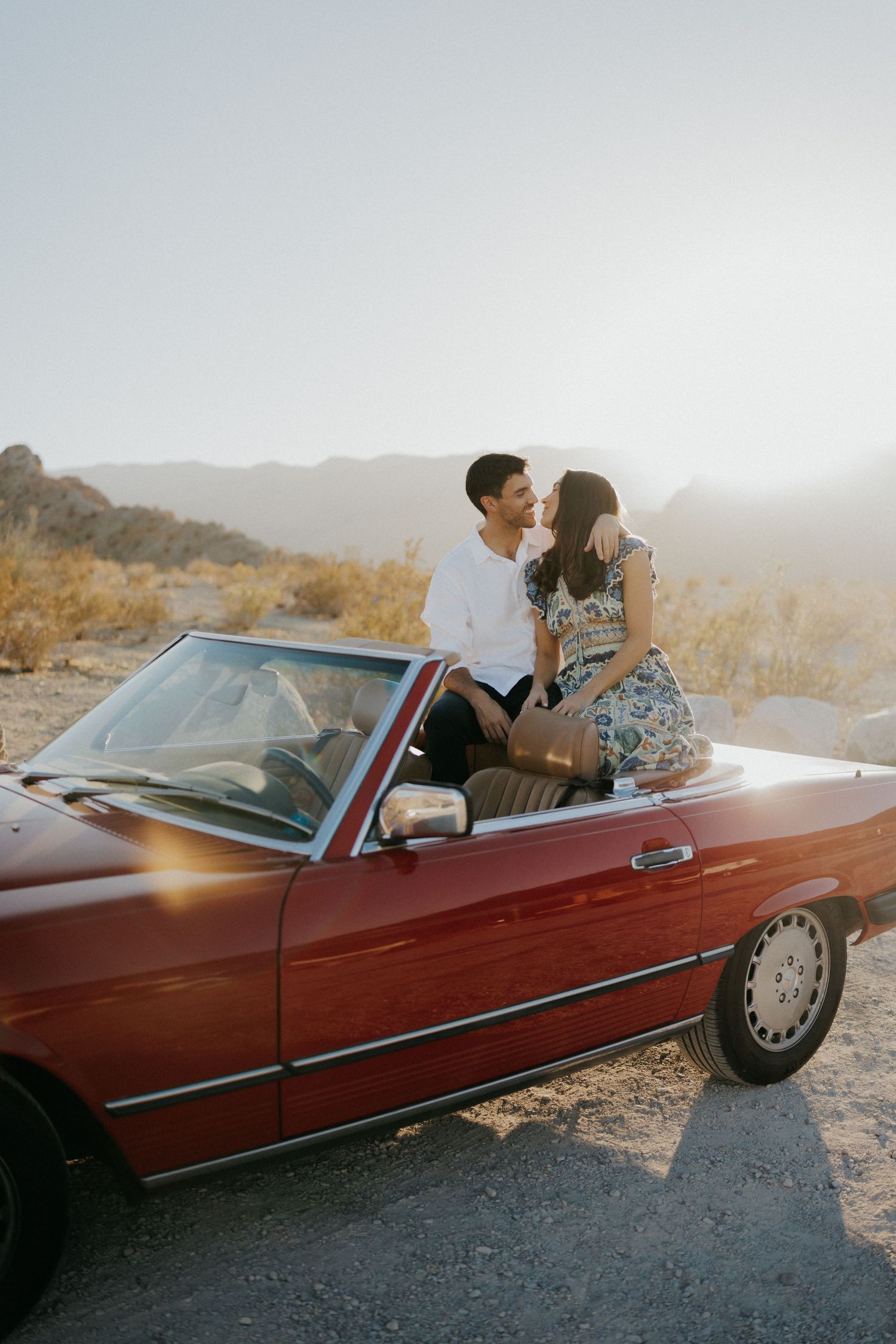 Couple sharing romantic moments in a vintage red convertible car parked in a desert landscape at golden hour.