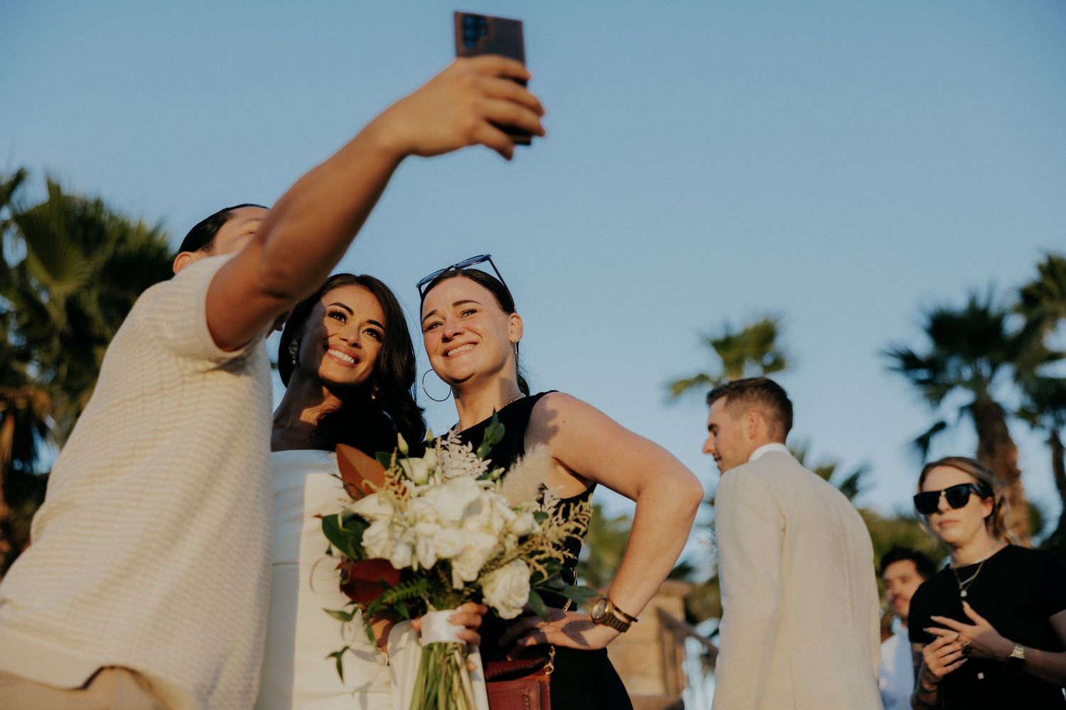 A couple shares their first dance at a sunset wedding celebration surrounded by palm trees and joyful guests.