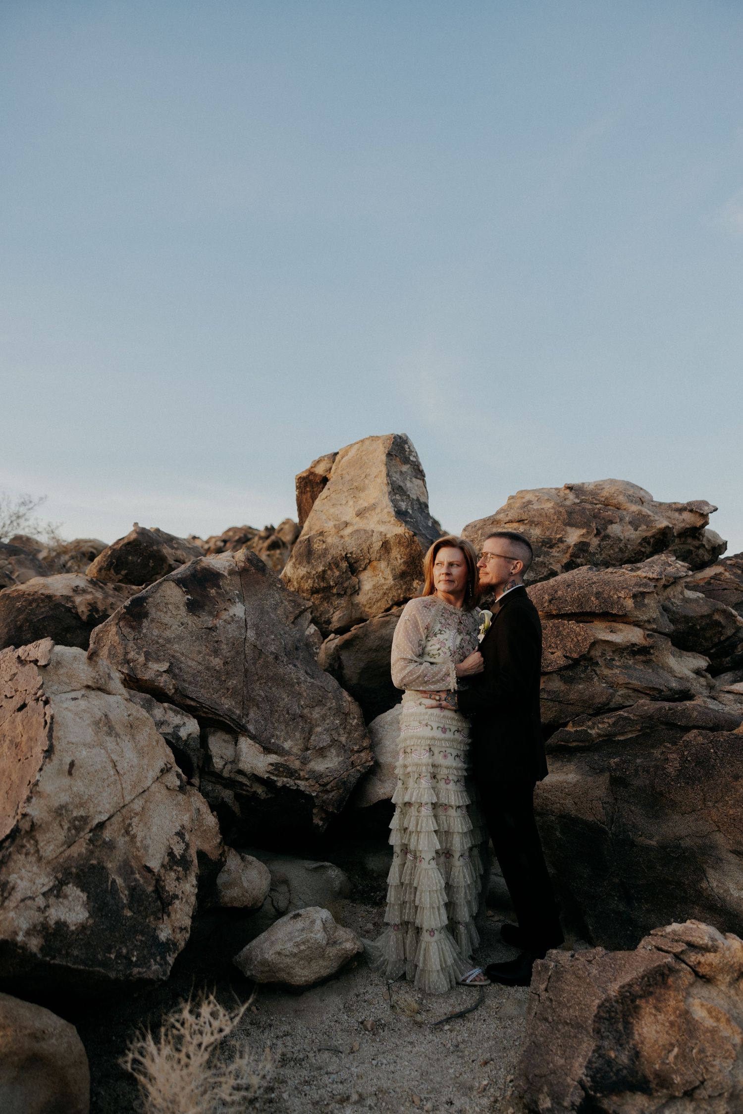 A couple in wedding attire poses together against dramatic desert rock formations at sunset.