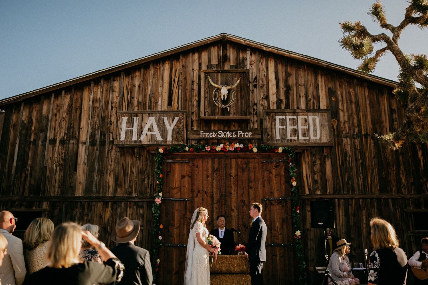 A rustic wooden barn with HAY FEED lettering hosts a gathering beneath antler decor and warm lighting.