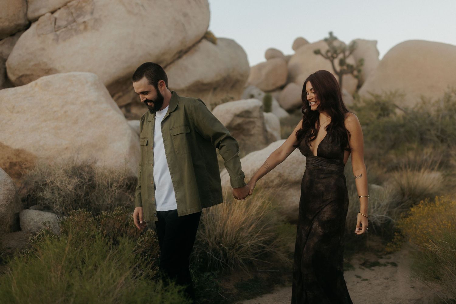 Couple holds hands walking among desert boulders, woman in black dress, man in olive jacket, Joshua Tree landscape.