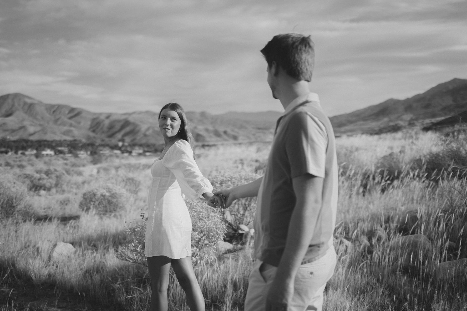Black and white photo of a couple holding hands in a scenic mountain field, woman in white dress looking back smiling.