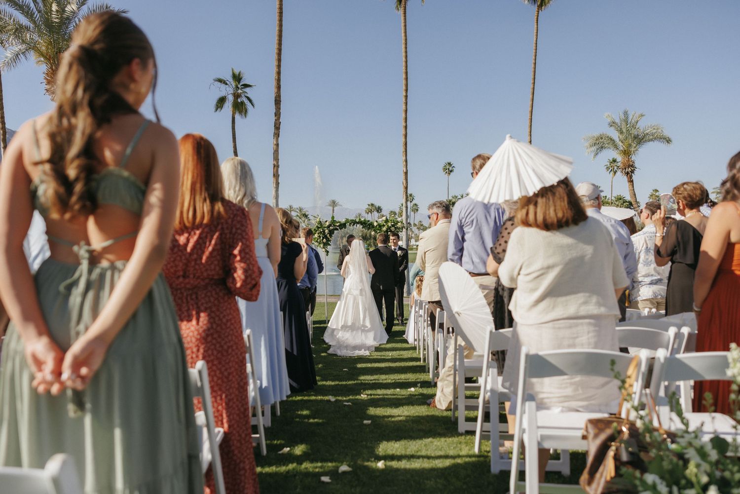 Outdoor wedding ceremony with guests seated in white chairs along a lush green aisle lined with palm trees.