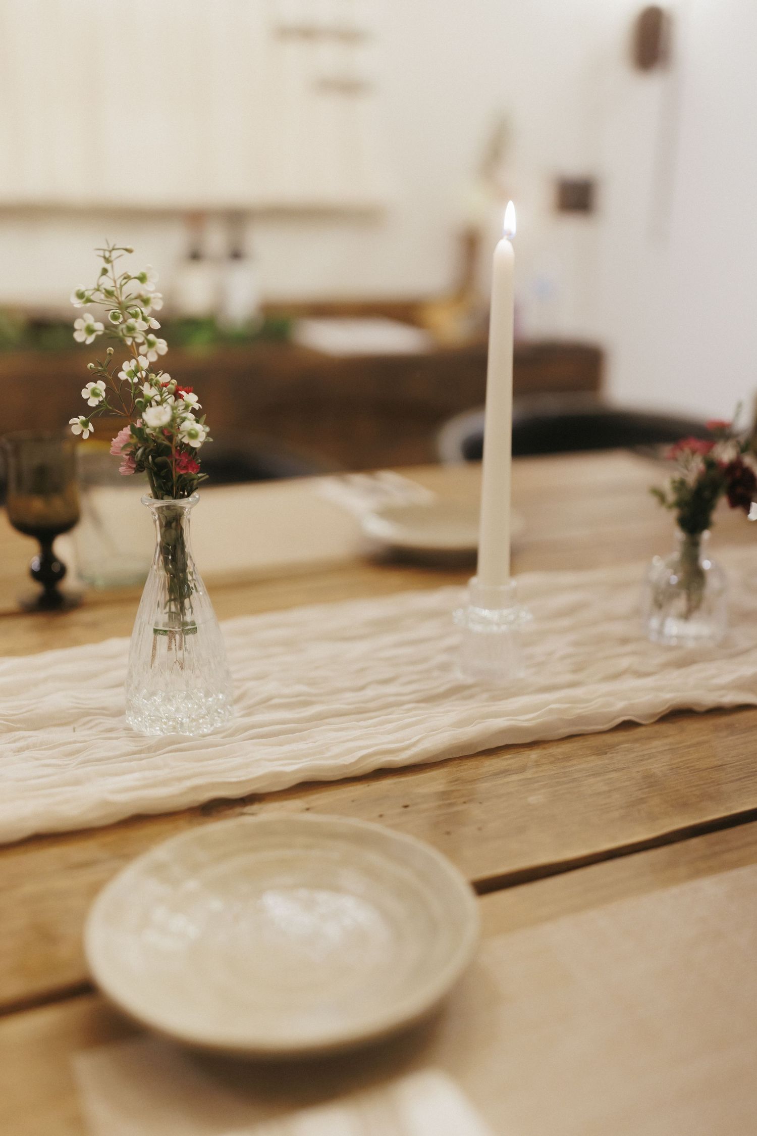 Rustic wooden wedding reception table with candle, small floral vases, and linen table runner.