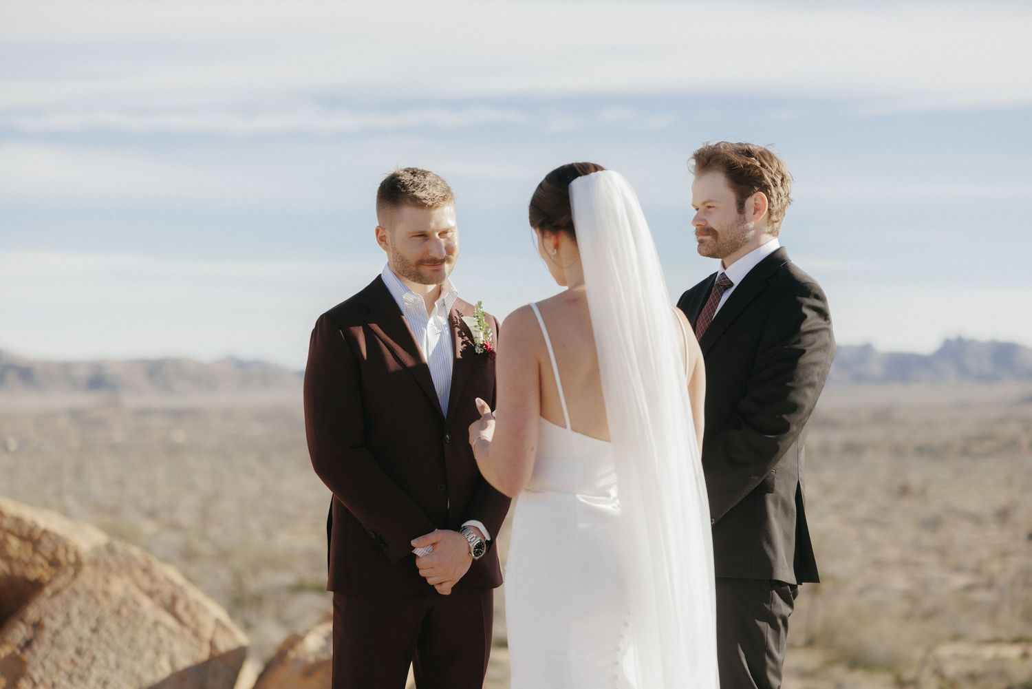 Couple exchanging vows with officiant in desert setting under cloudy sky.