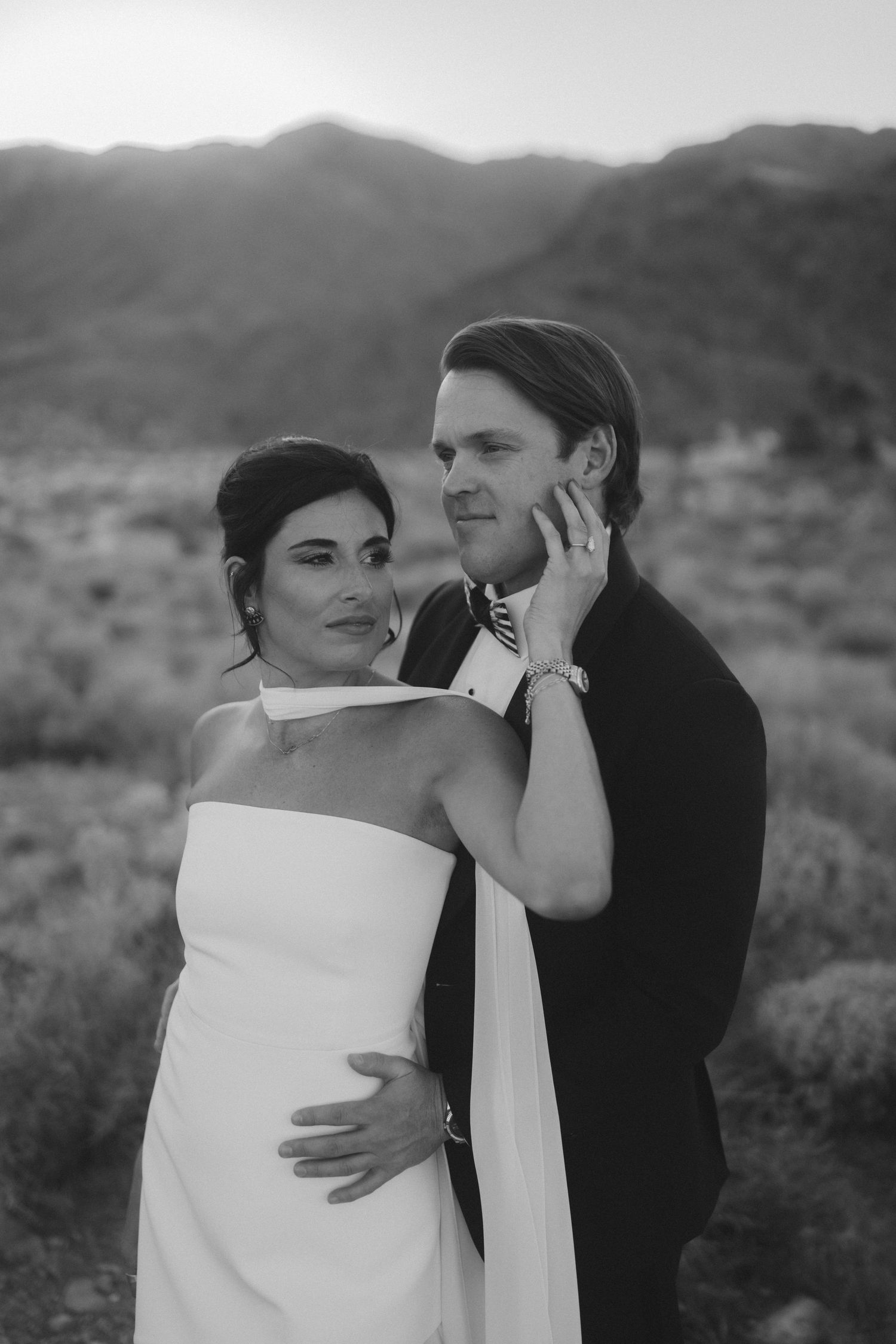 Black and white portrait of bride and groom embracing in desert landscape with mountains in background.