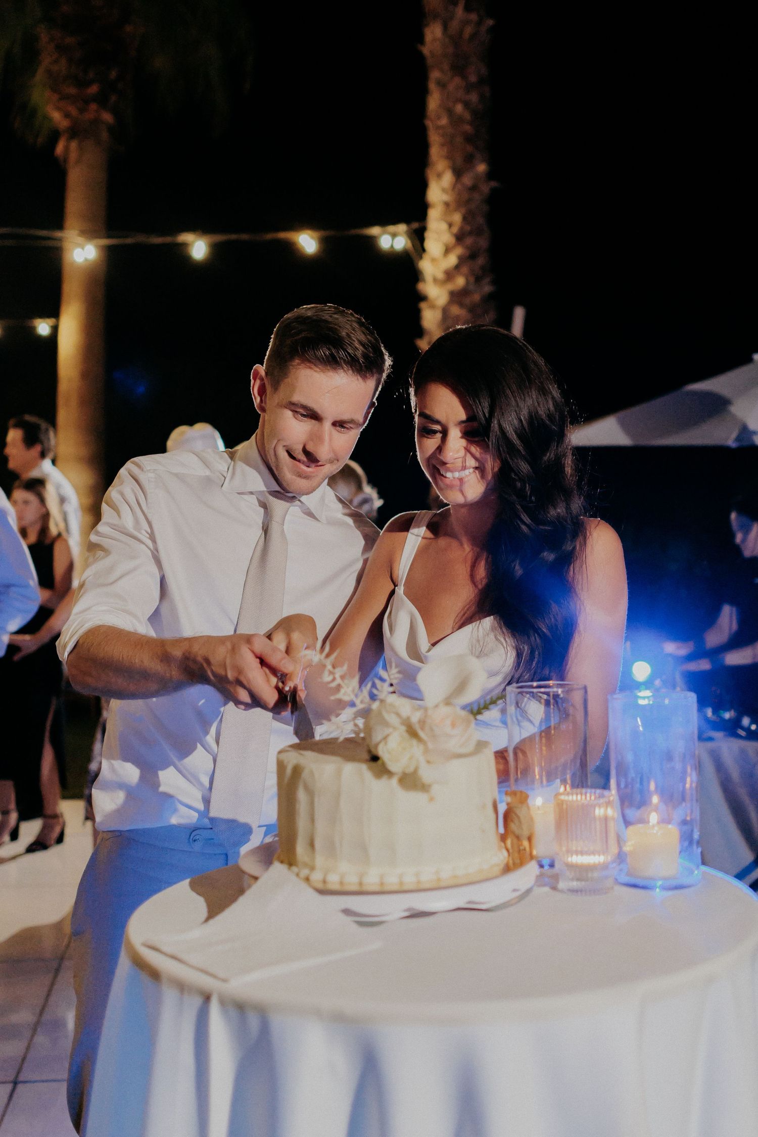 A couple cuts their wedding cake together at a dimly lit evening reception with string lights overhead.