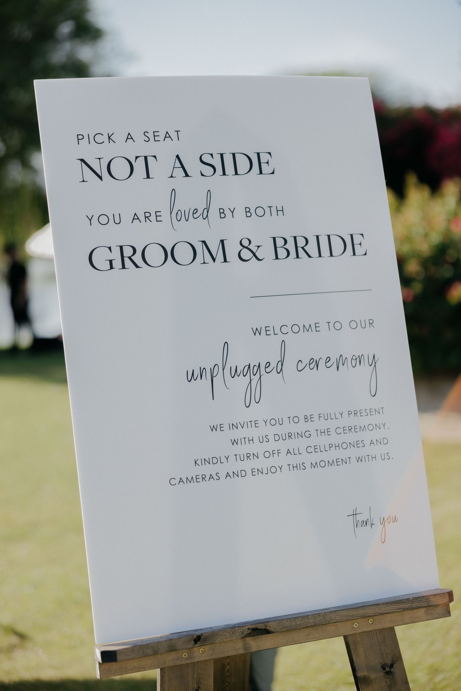 Wedding ceremony sign on wooden easel displaying 'Pick a seat not a side - you are loved by both Groom & Bride' outdoors.
