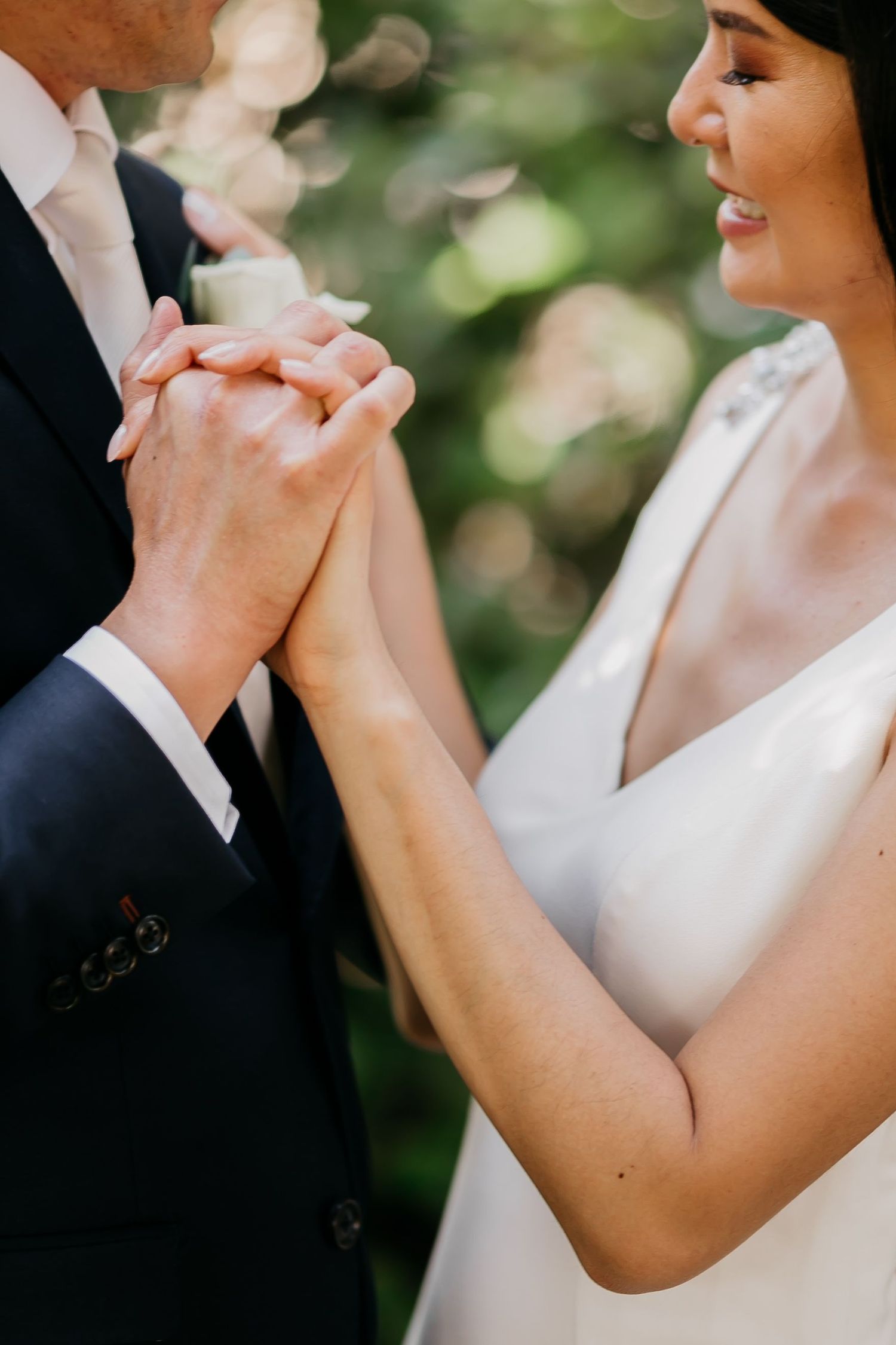 Closeup of wedding couple holding hands tenderly outdoors with soft bokeh light and greenery in background.