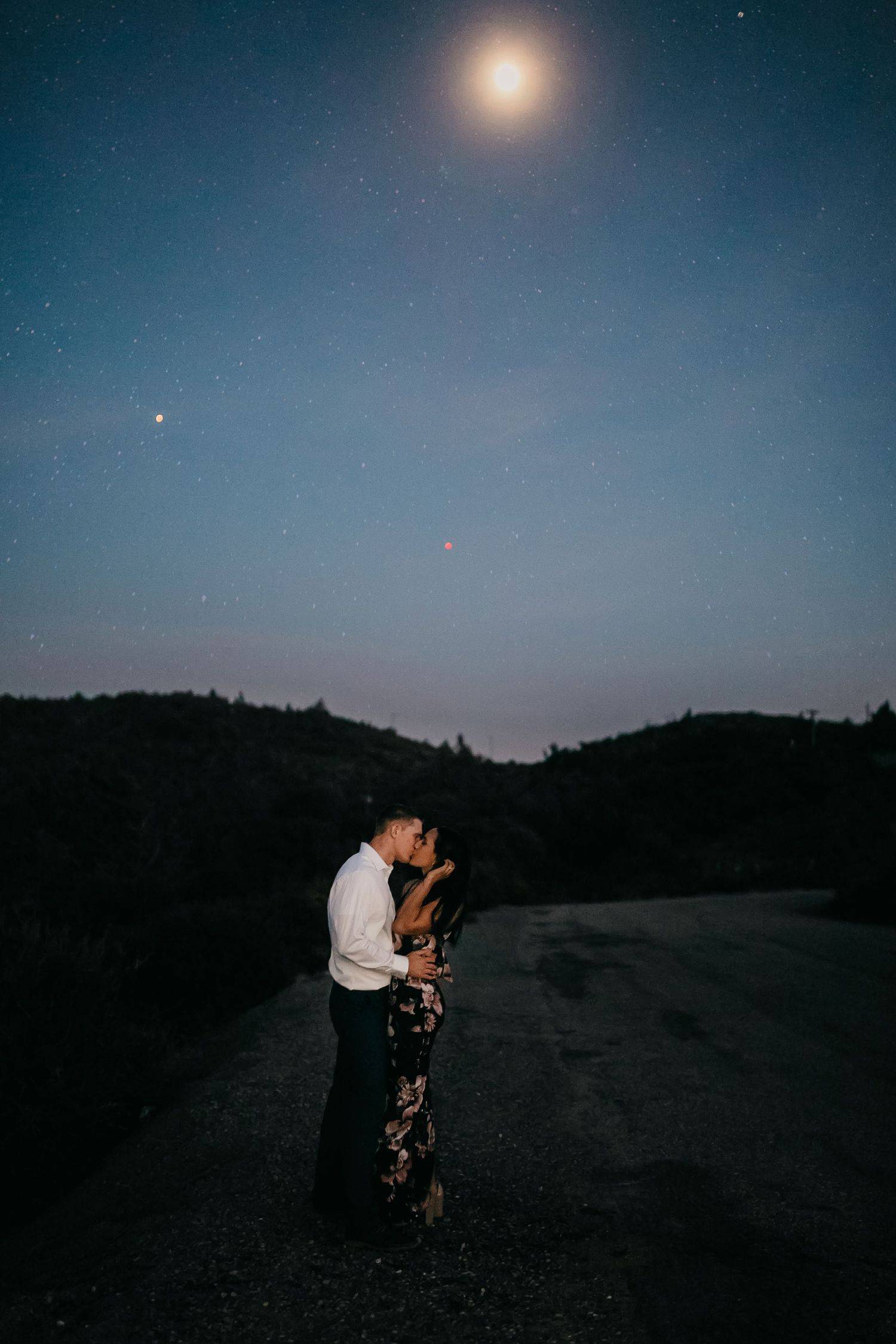 A romantic nighttime photoshoot beneath a bright full moon showing silhouettes against a dark hillside landscape.