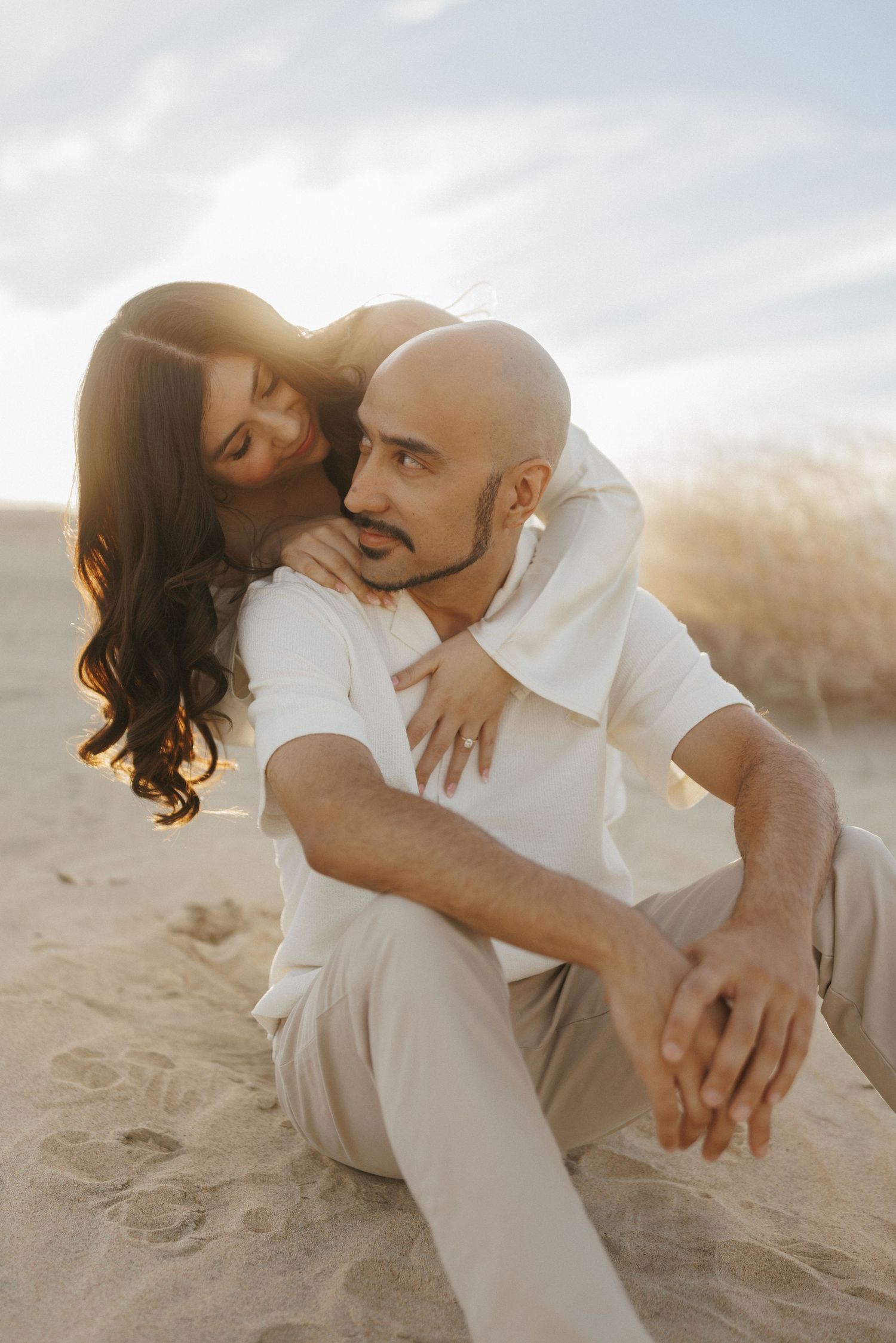 A bald man and a woman with long dark hair share a romantic embrace on a sandy beach at golden hour.