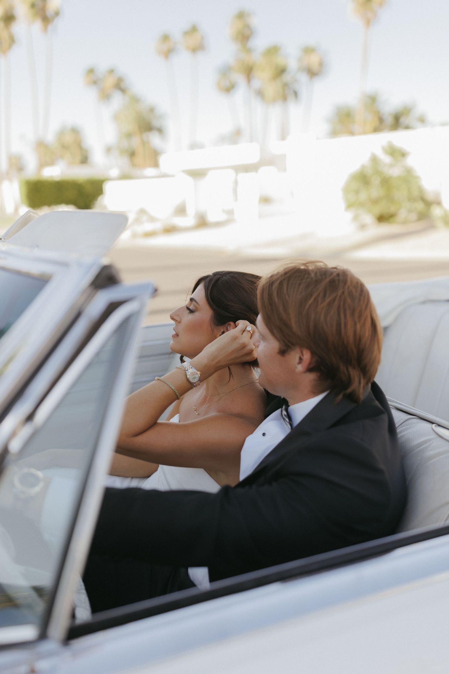 A bride and groom in formal attire ride in a white convertible car surrounded by palm trees.