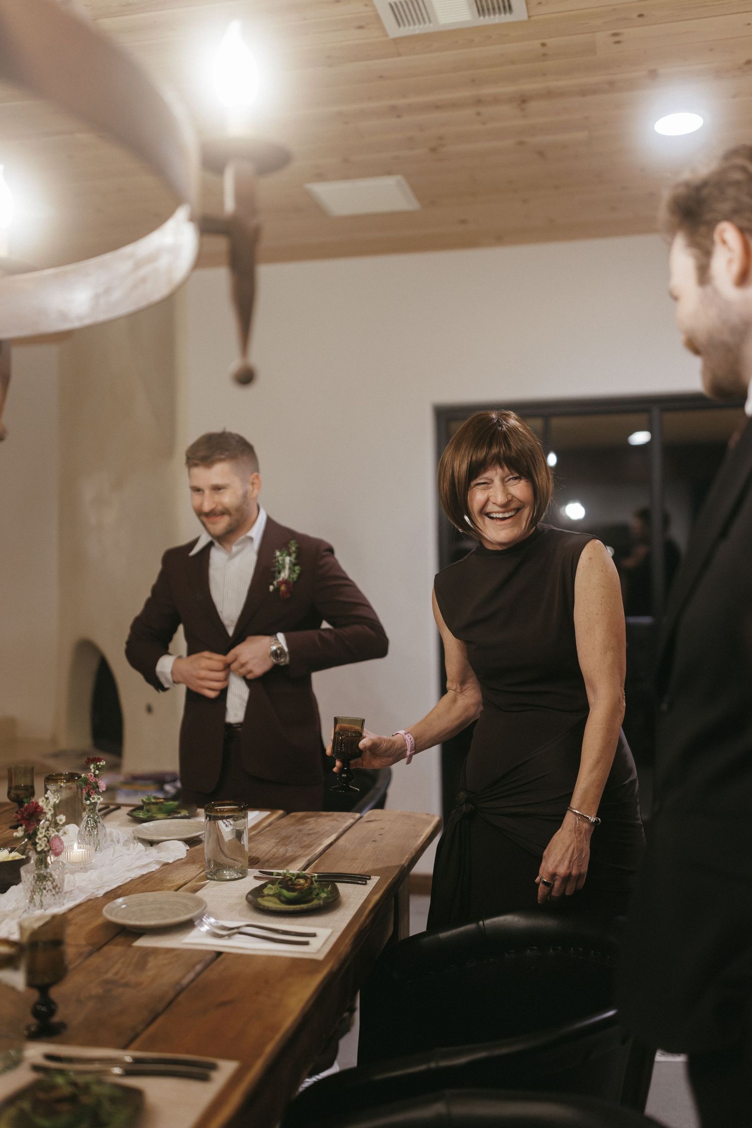 Elegant dinner party with guests in formal attire gathered around a rustic wooden table in a warmly lit venue.
