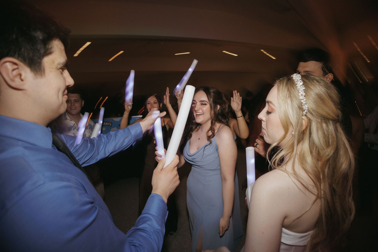 Bride and guests dancing with glowing foam sticks at lively indoor wedding reception party.