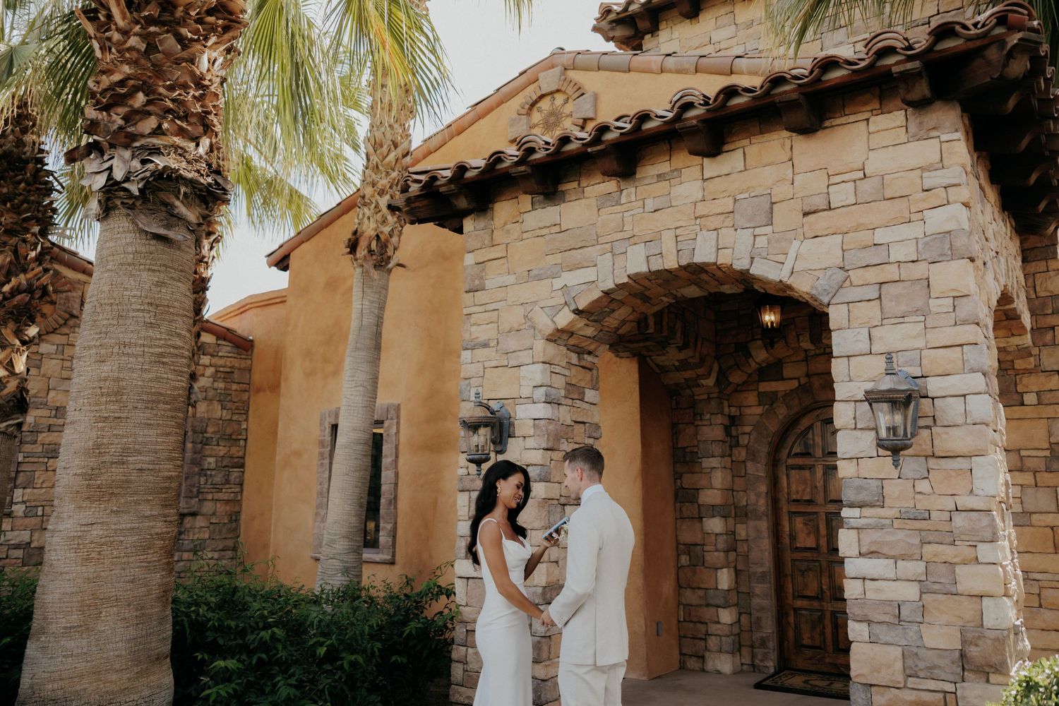 Rustic stone archway entrance with decorative light fixture and palm trees against a beige Mediterranean style building.